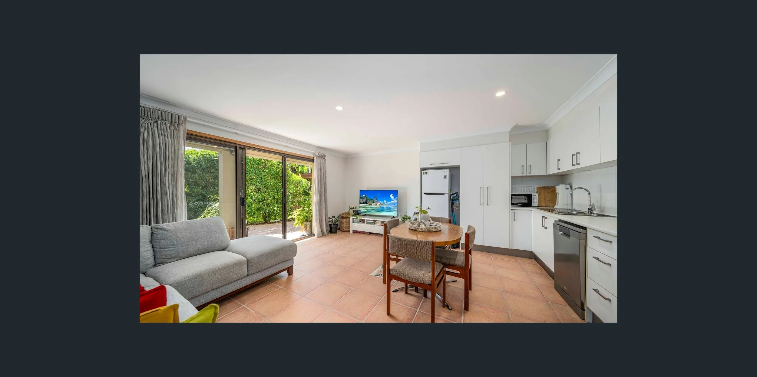 Living room and kitchen with sliding glass door leading to outdoor greenery, gray sofa, round dining table with four chairs, white cabinetry, appliances, and a TV.