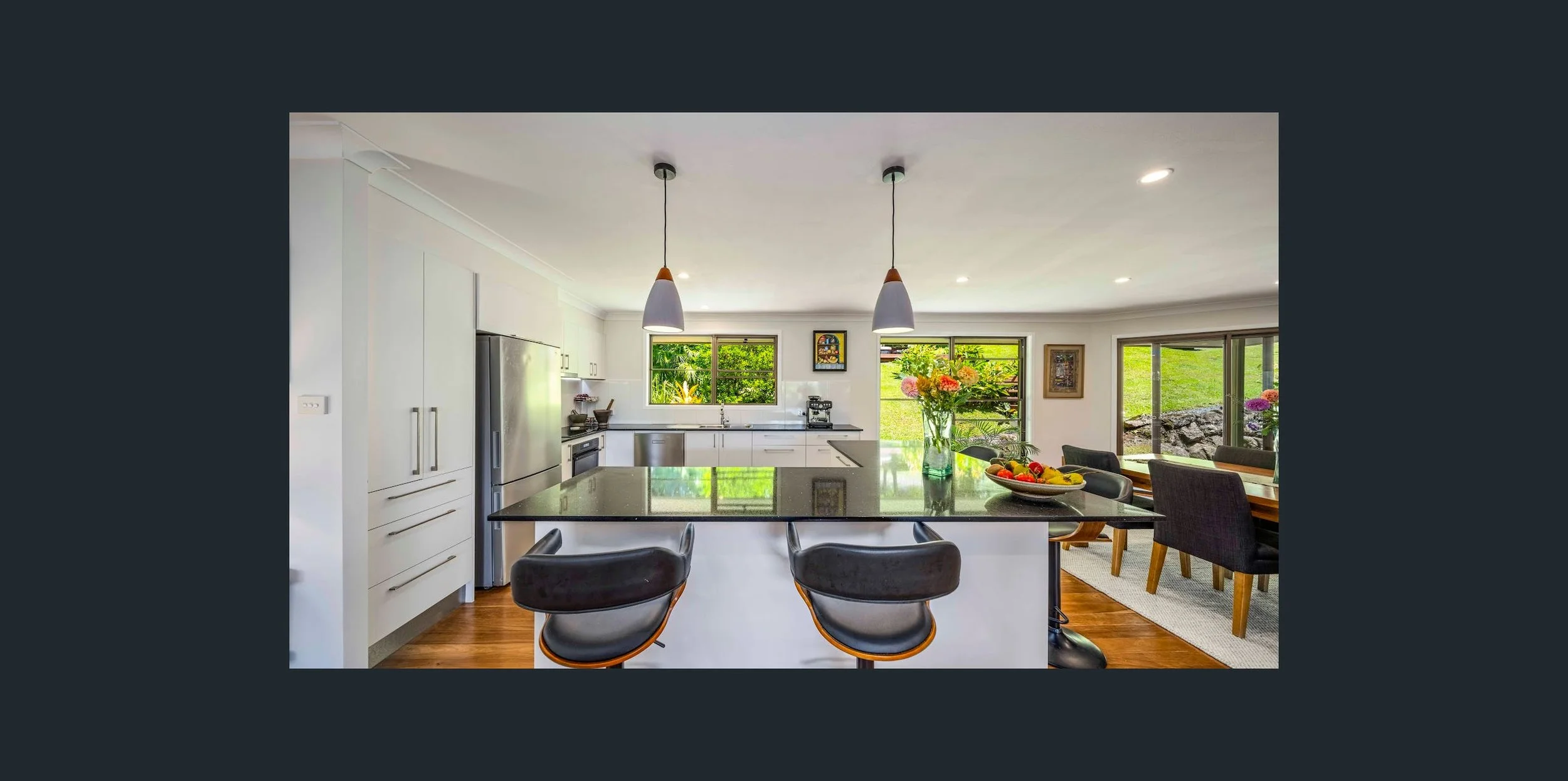 Modern kitchen with white cabinets, stainless steel refrigerator, black countertops, and a central island with bar stools. Large windows overlook a lush green yard, and there is a dining table with chairs and a vase of flowers.
