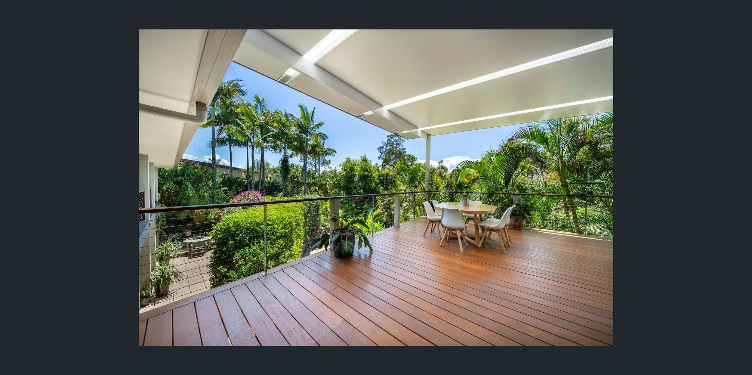 Spacious outdoor balcony with wooden flooring, surrounded by lush green tropical plants, including palm trees; features a round table with four chairs and potted plants, under a white ceiling with integrated lighting, overlooking a garden area.