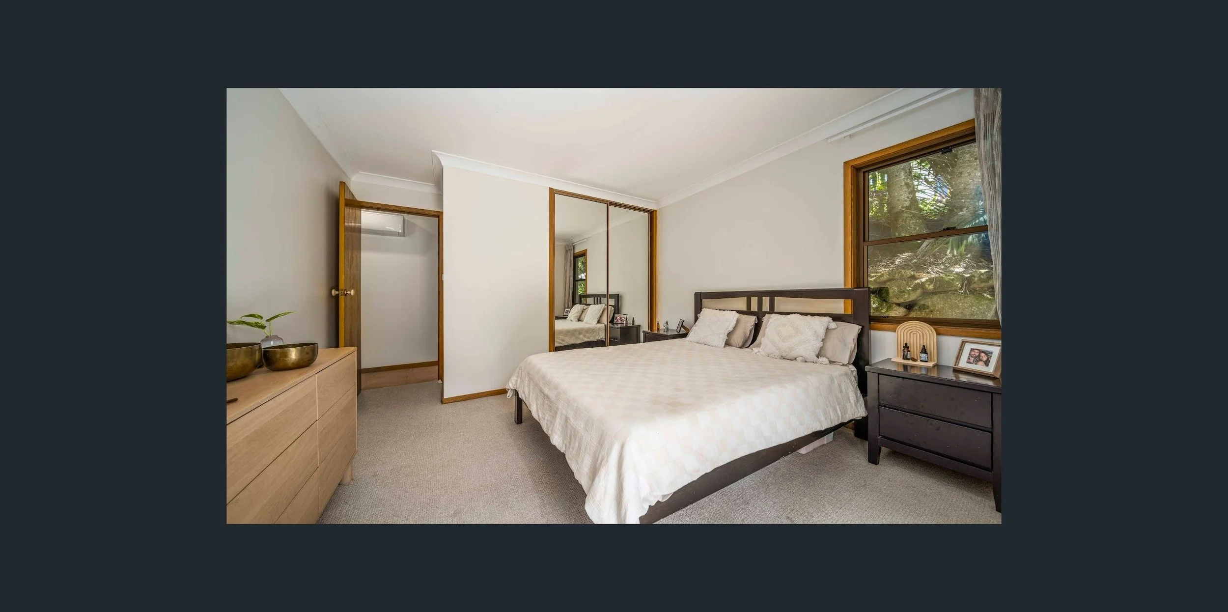 A bedroom with a large bed, a nightstand with framed photo and bottles, a window showing trees outside, and a dresser with decorative bowls and plants.