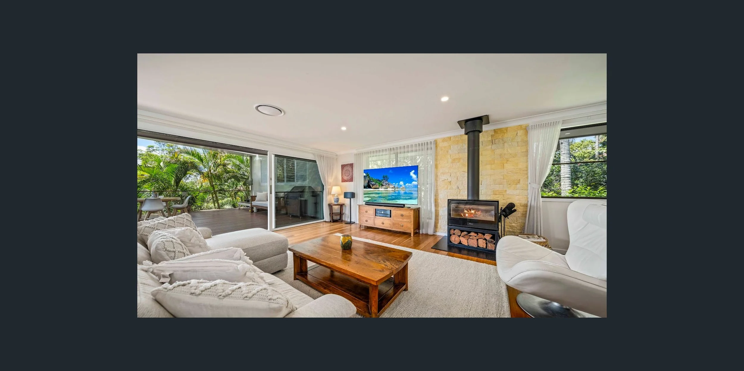 Living room with a view of lush greenery, featuring a fireplace, a large TV, white and wooden furniture, and sliding glass doors opening to a balcony with outdoor seating.