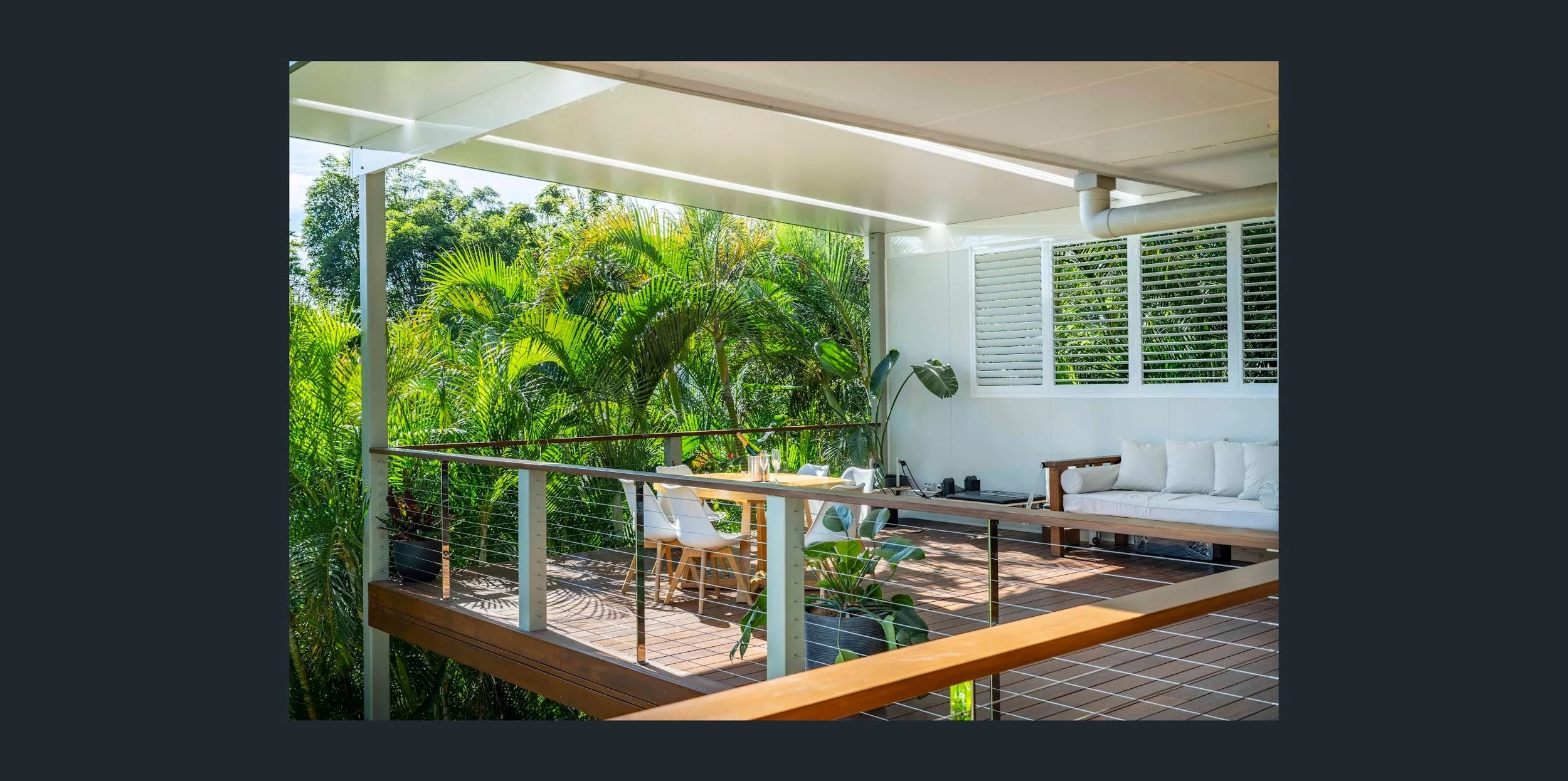 Balcony with white cushioned sofa, wooden table, and chairs amid lush green foliage outside.