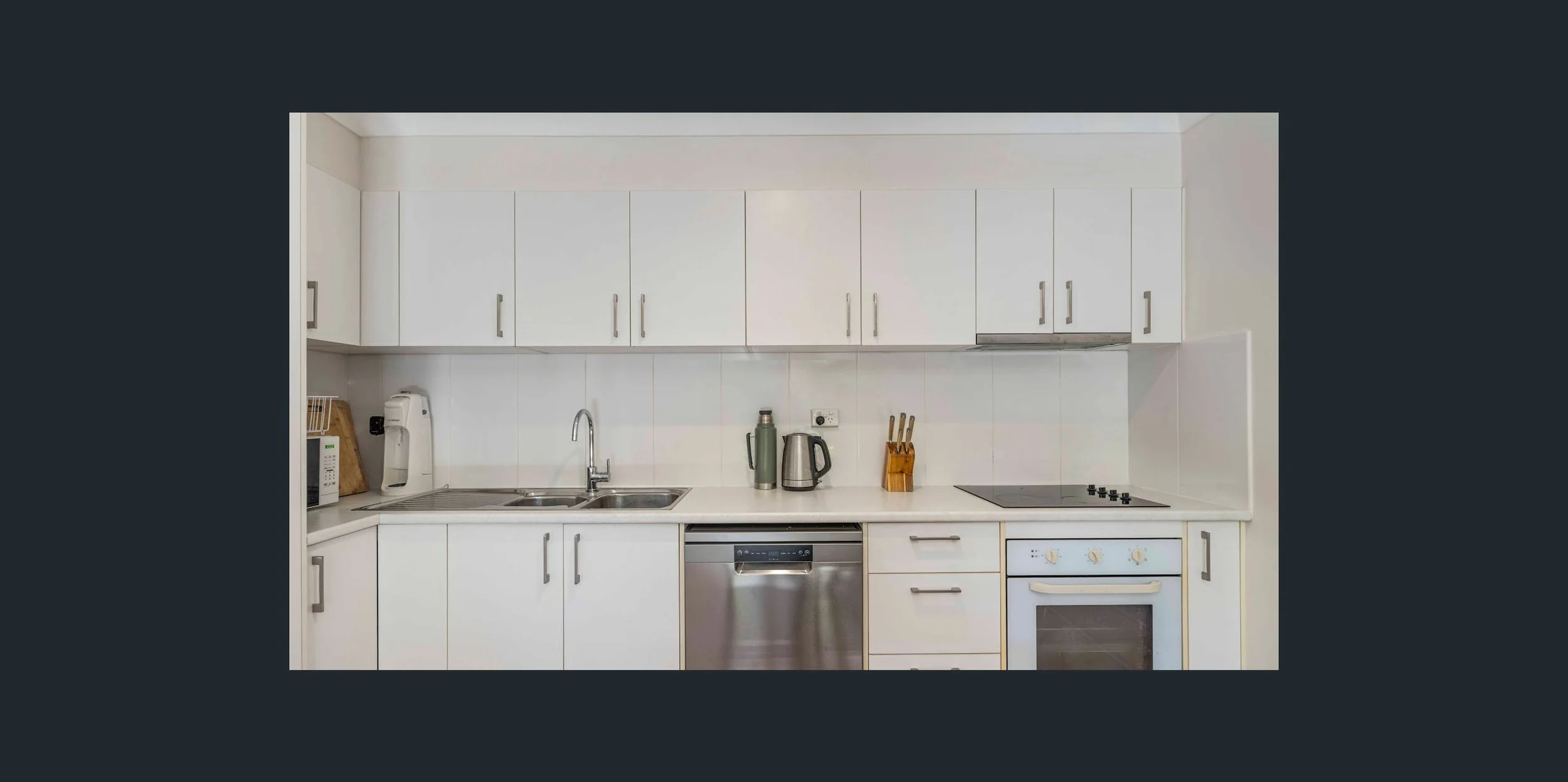 A modern kitchen with white cabinets, a stainless steel dishwasher, a sink with a silver faucet, a microwave, coffee machine, kettle, and knife block on the countertop.