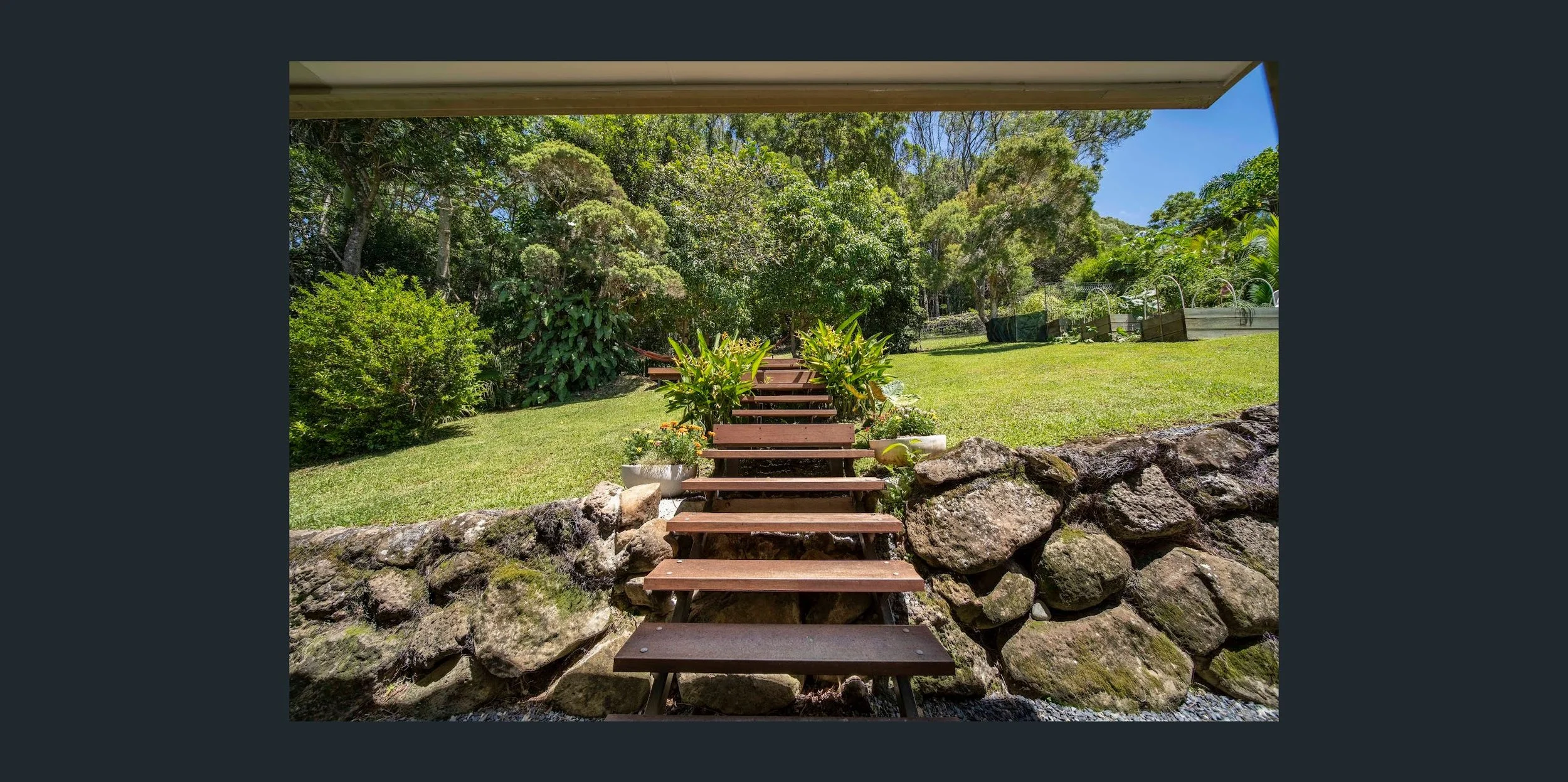 Wooden steps leading up a stone wall into a lush green garden with trees and plants, under a clear blue sky.