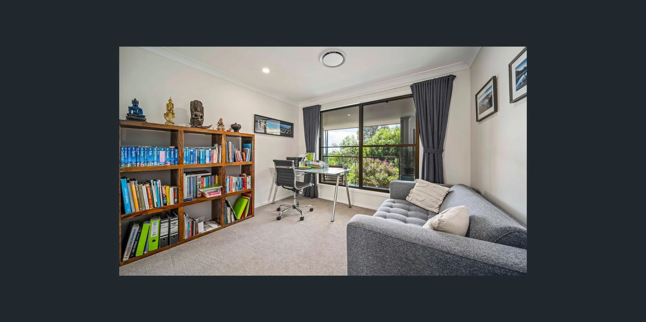 A cozy, well-lit home office with a view of greenery outside. It features a gray sofa, a wooden bookshelf filled with books, decorative statues, and binders, a black office chair, a desk with a computer and office supplies, and framed photos on the w