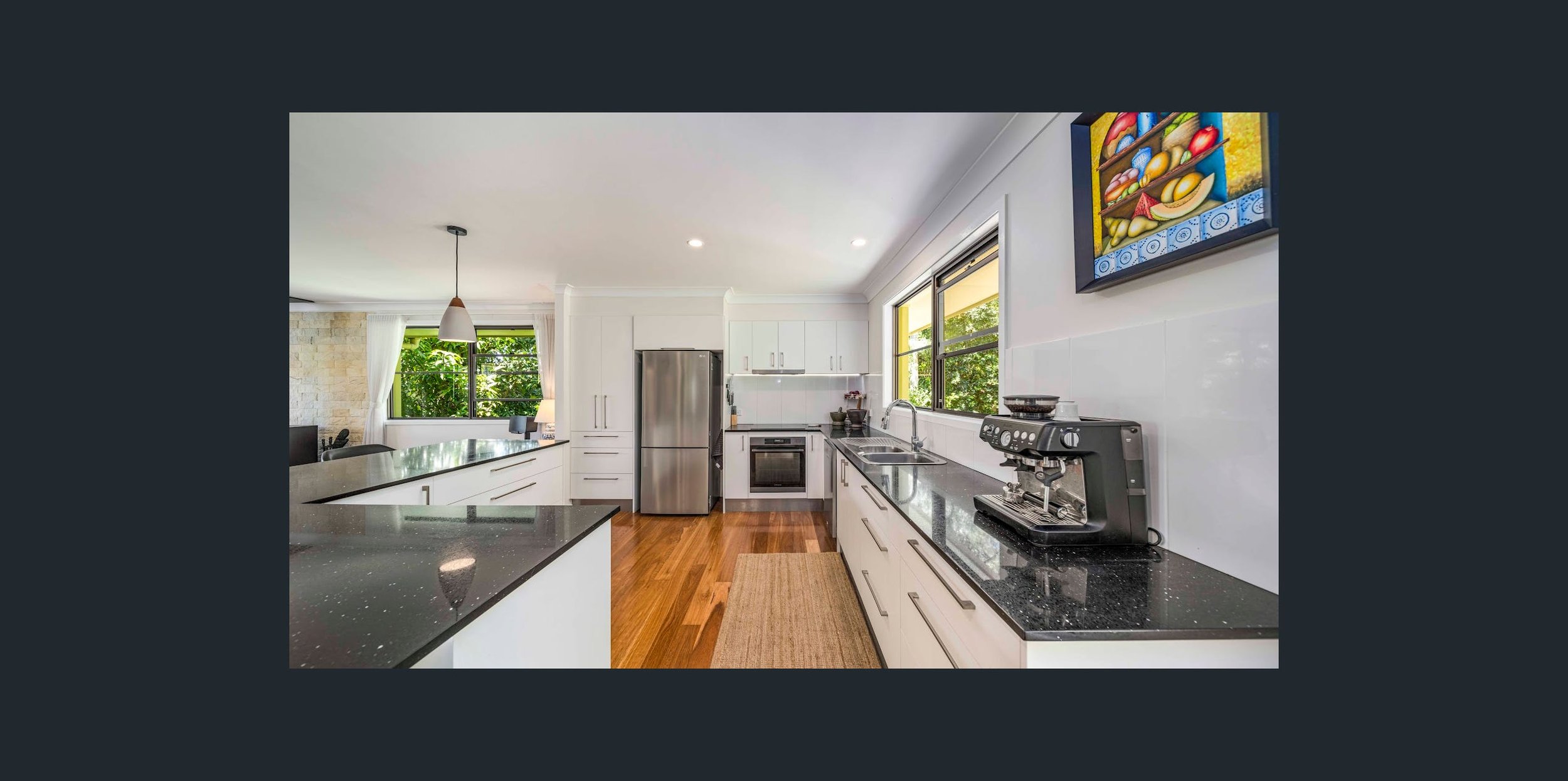 Modern kitchen with black countertops, white cabinets, stainless steel appliances, and hardwood floors. Features a wall-mounted colorful painting, a coffee machine, and large windows with greenery outside.