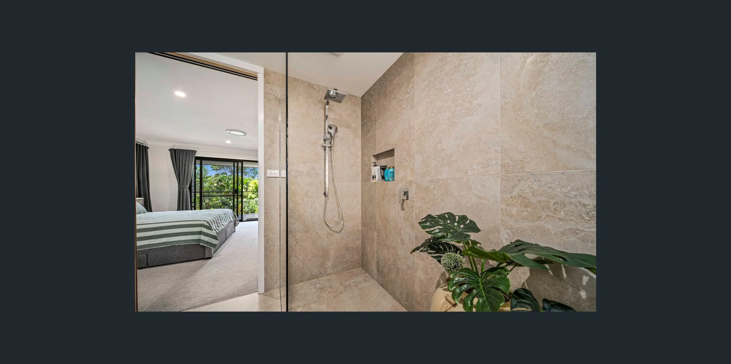 Modern bedroom with a large window, view of greenery, and a glass-walled shower with beige tiles, a built-in shelf, and a potted plant.