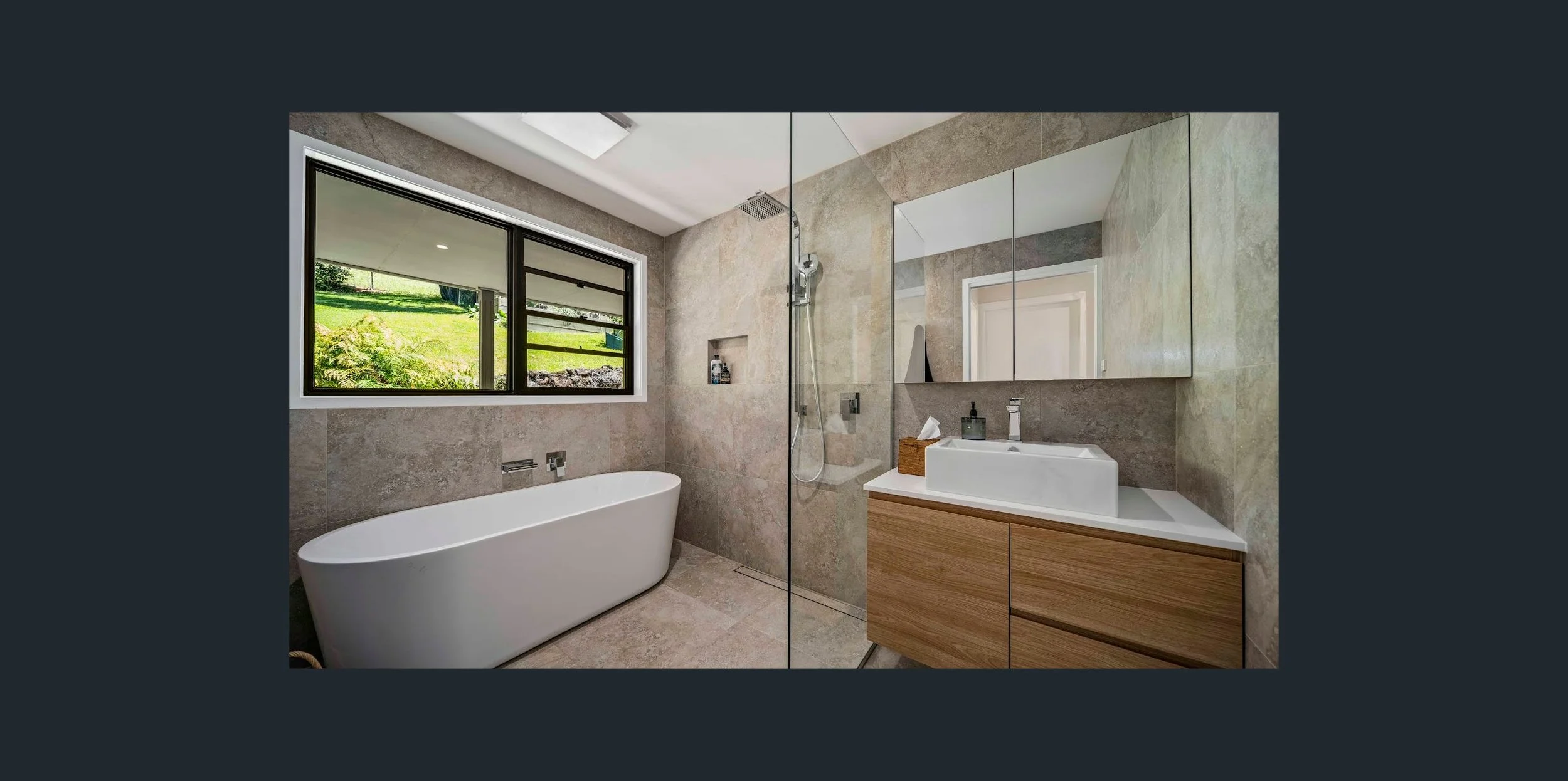 Modern bathroom with a bathtub, large window showing green outdoor scenery, walk-in shower with a glass partition, and a wooden vanity with a white sink and mirror cabinets.