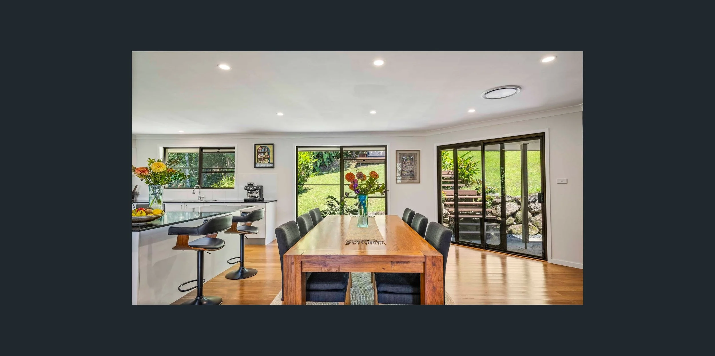 A bright dining area with a large wooden table, black chairs, and a view of a green backyard through sliding glass doors and windows. Decorative flowers are on the table and counter.
