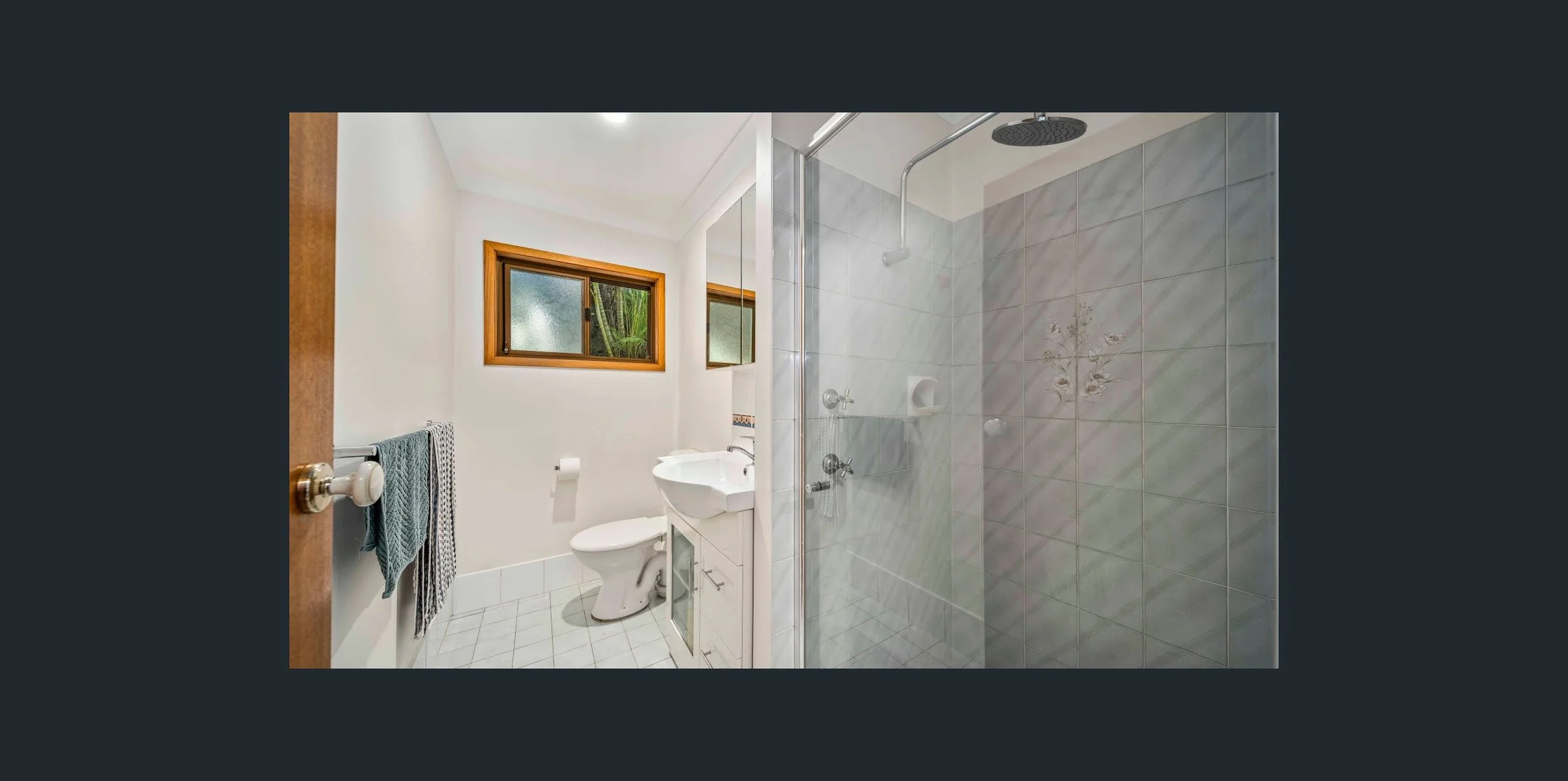 Small bathroom with white walls, two small windows with wooden frames, a toilet, a small sink, and a glass-encased shower with tiled walls.