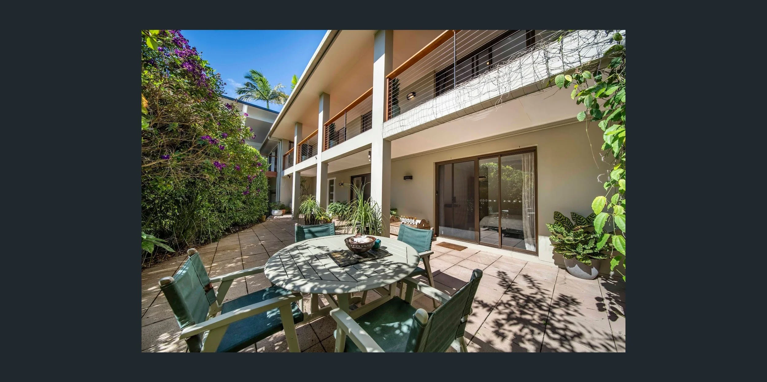 Outdoor patio area with a round table and six chairs, surrounded by lush greenery and potted plants, attached to a two-story building with sliding glass doors and balcony.