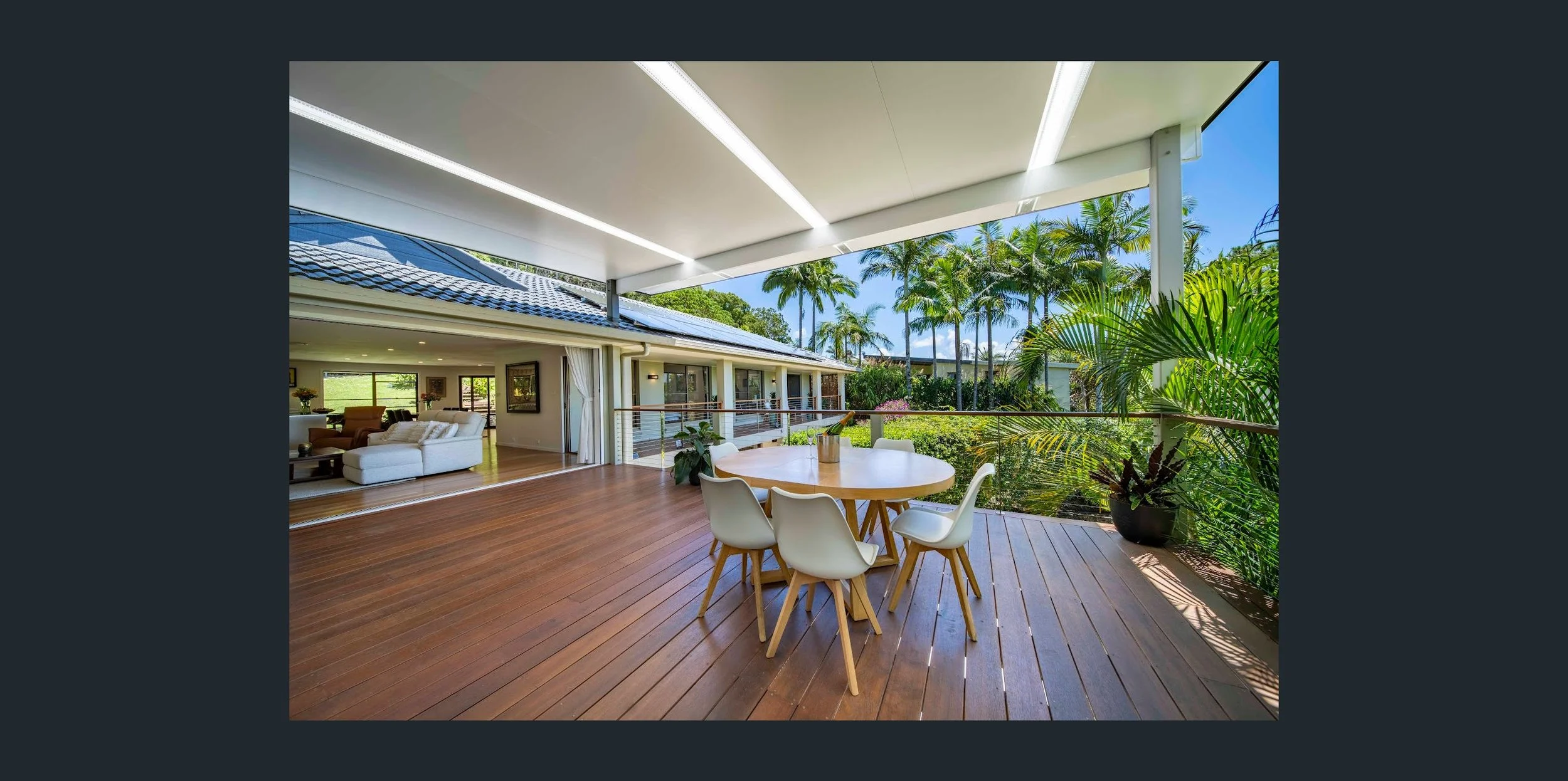 Open outdoor patio with wooden flooring, a round dining table with four white chairs, potted plants, lush green trees, and a view of a house interior with a white sofa and windows.