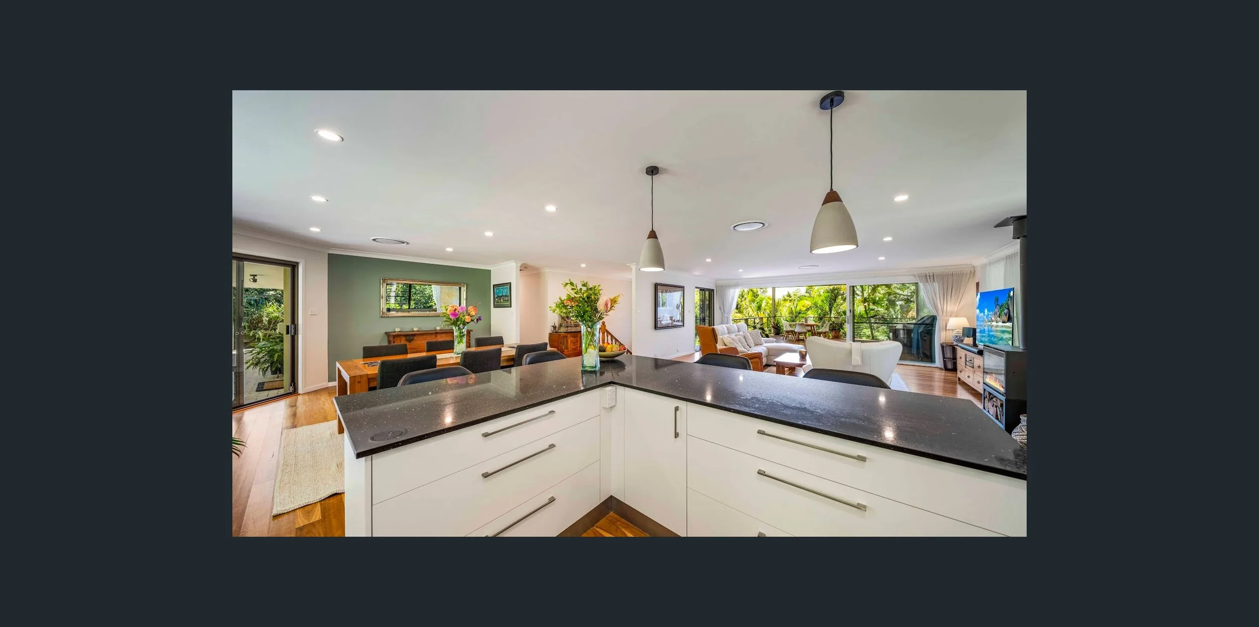 Open-concept living room and kitchen with large windows, hardwood floors, modern furniture, and a view of greenery outside.