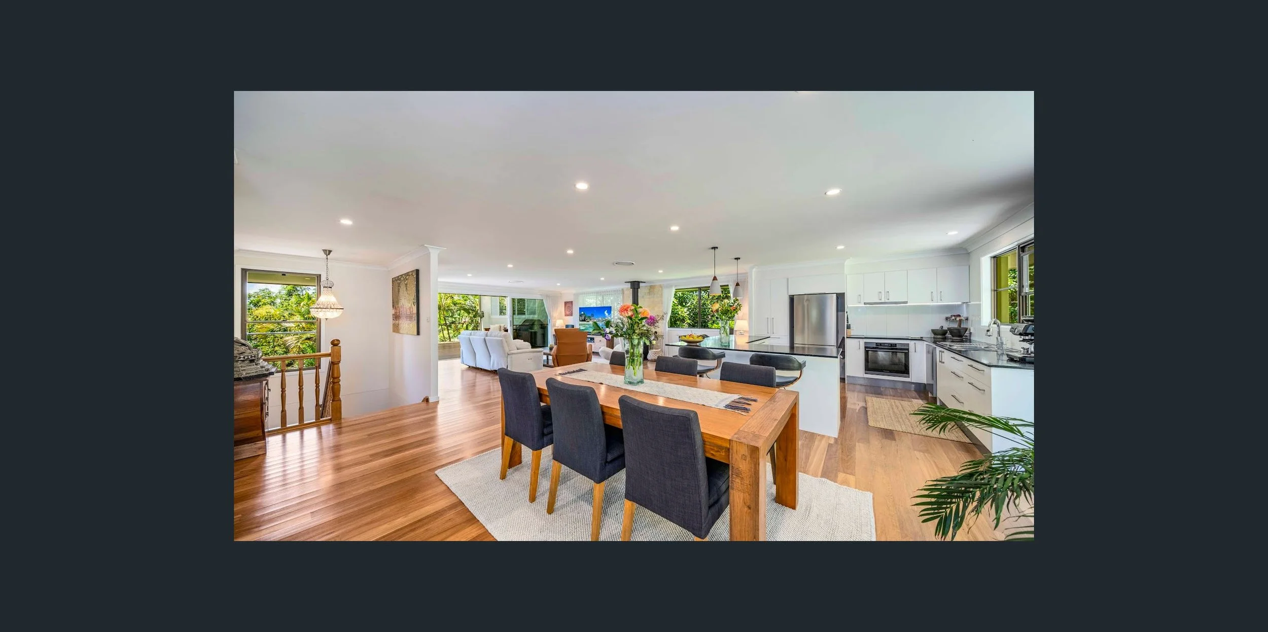 Open-concept living and kitchen area with hardwood floors, white cabinets, stainless steel appliances, and large windows showing greenery outside.