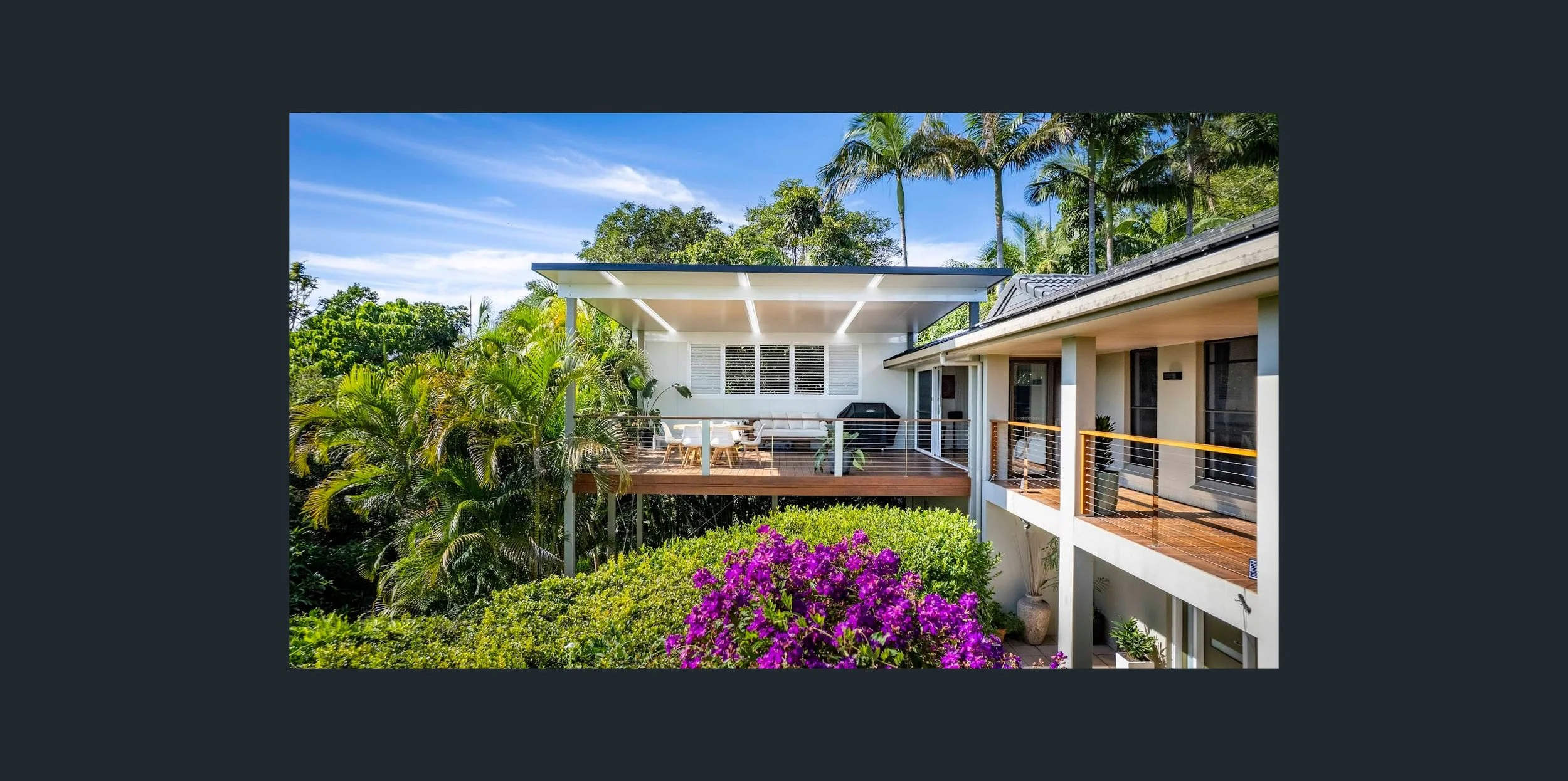 A modern house with white exterior and wooden balcony surrounded by lush tropical plants, including palm trees and vibrant purple flowers, under a clear blue sky.