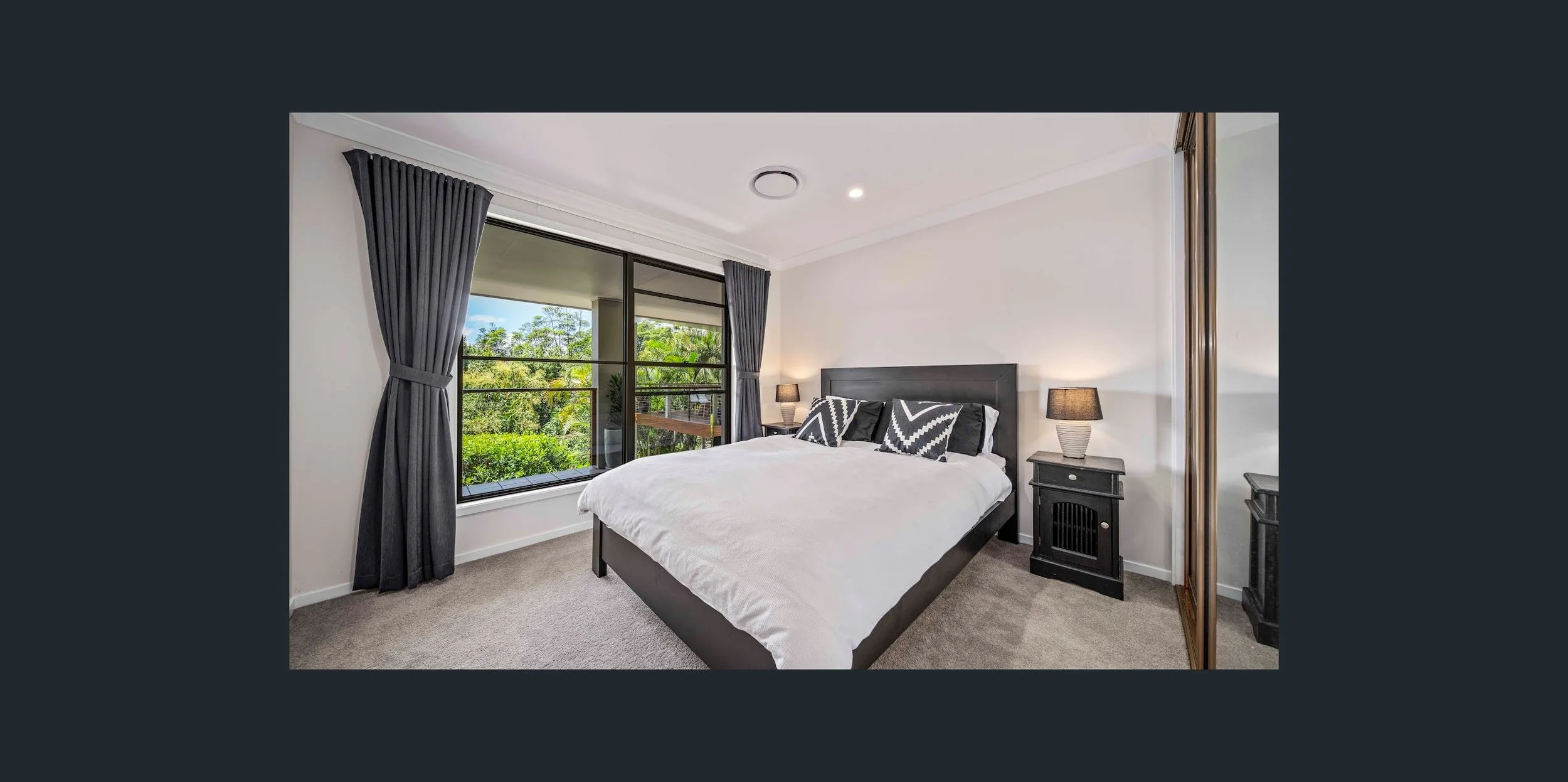 Modern bedroom with a large bed, black and white pillows, black nightstands with lamps, gray curtains, a large window showing greenery outside, and a closet with wooden doors.