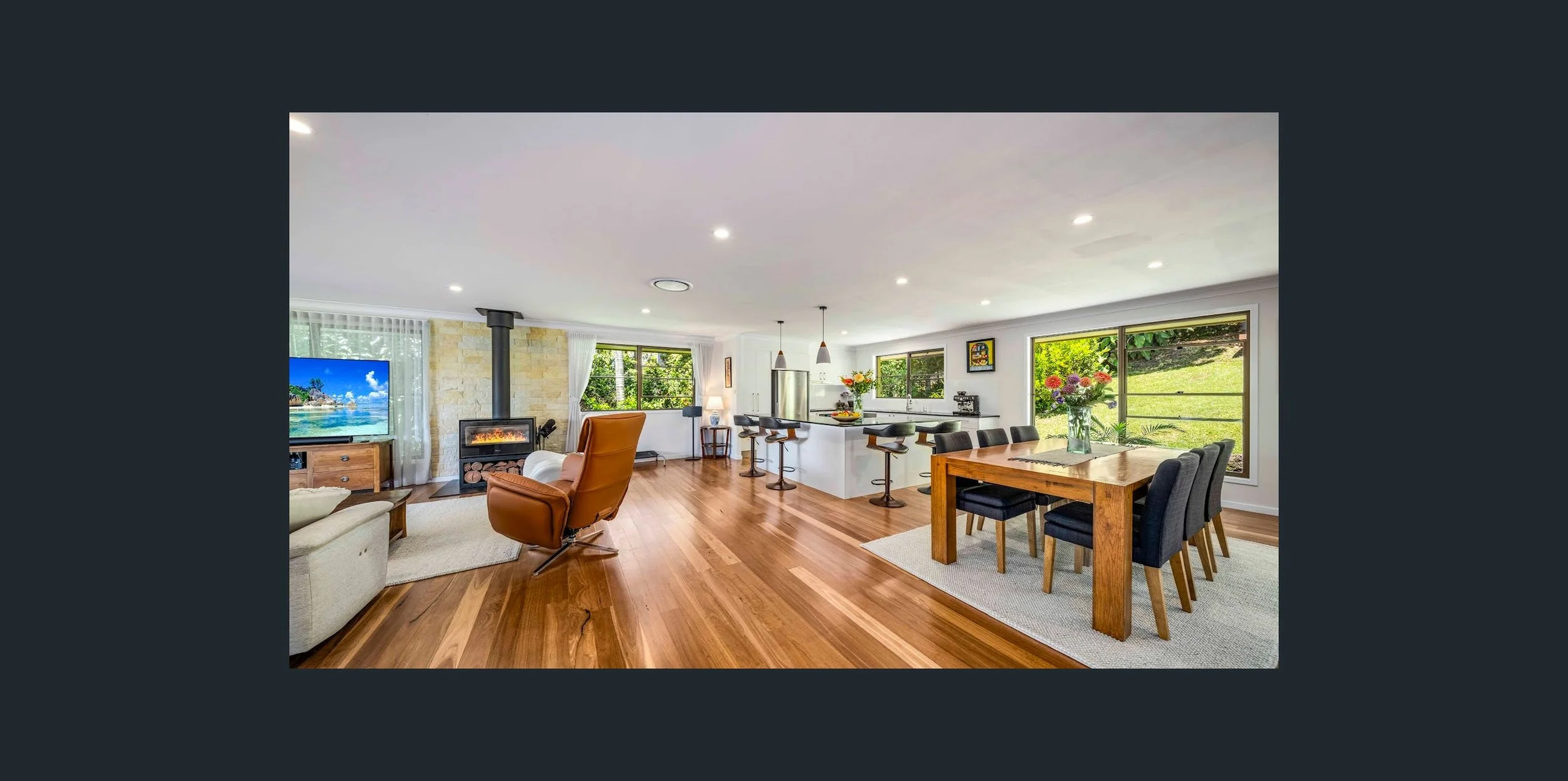 Open-concept living and dining area with hardwood floors, large windows, a fireplace, a TV, and a kitchen with bar seating and a dining table with chairs.