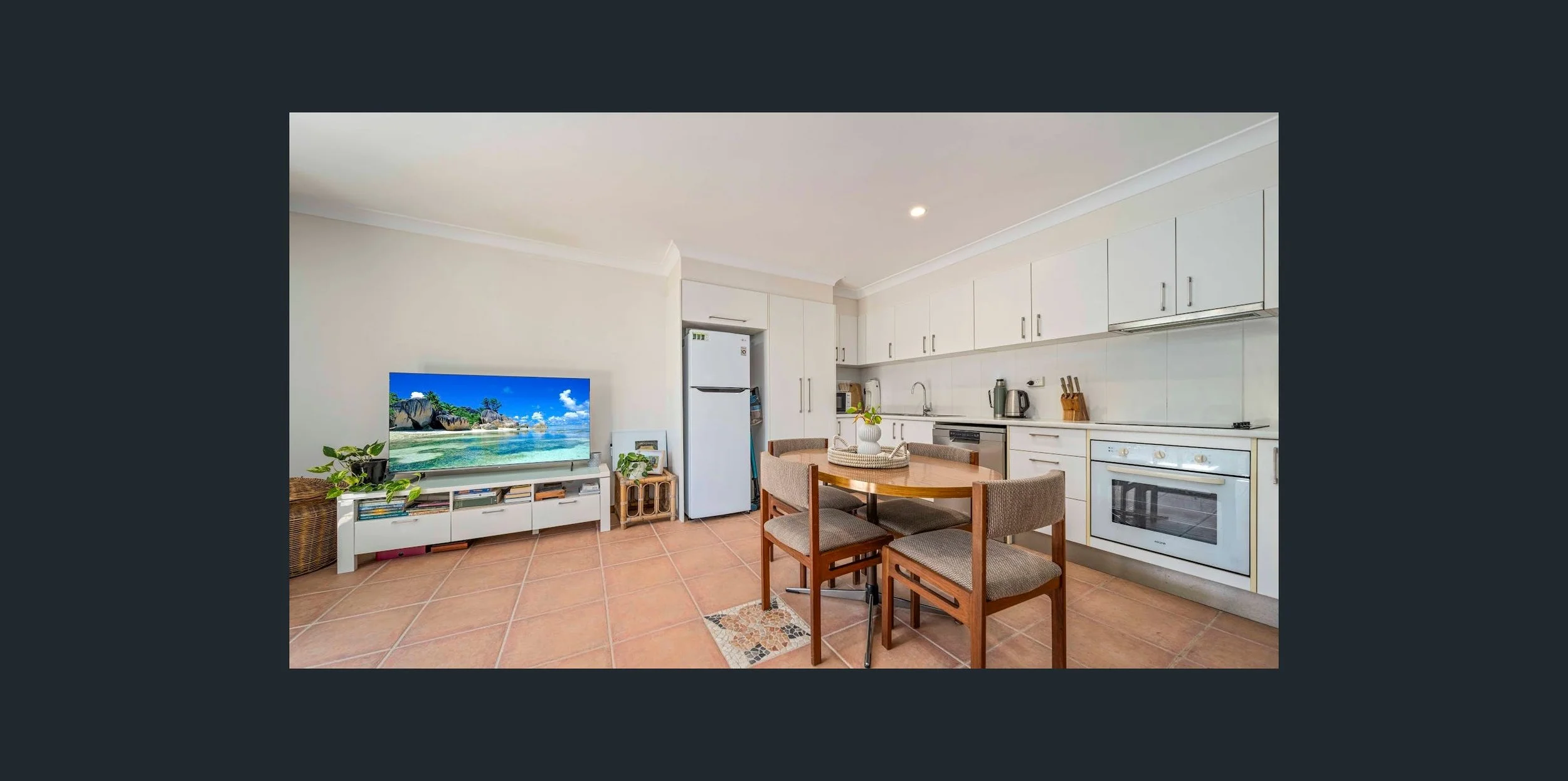 Open-plan kitchen and dining area with white cabinets, a white fridge, a round wooden table, four beige upholstered chairs, a TV on a white stand, and terracotta tiled flooring.