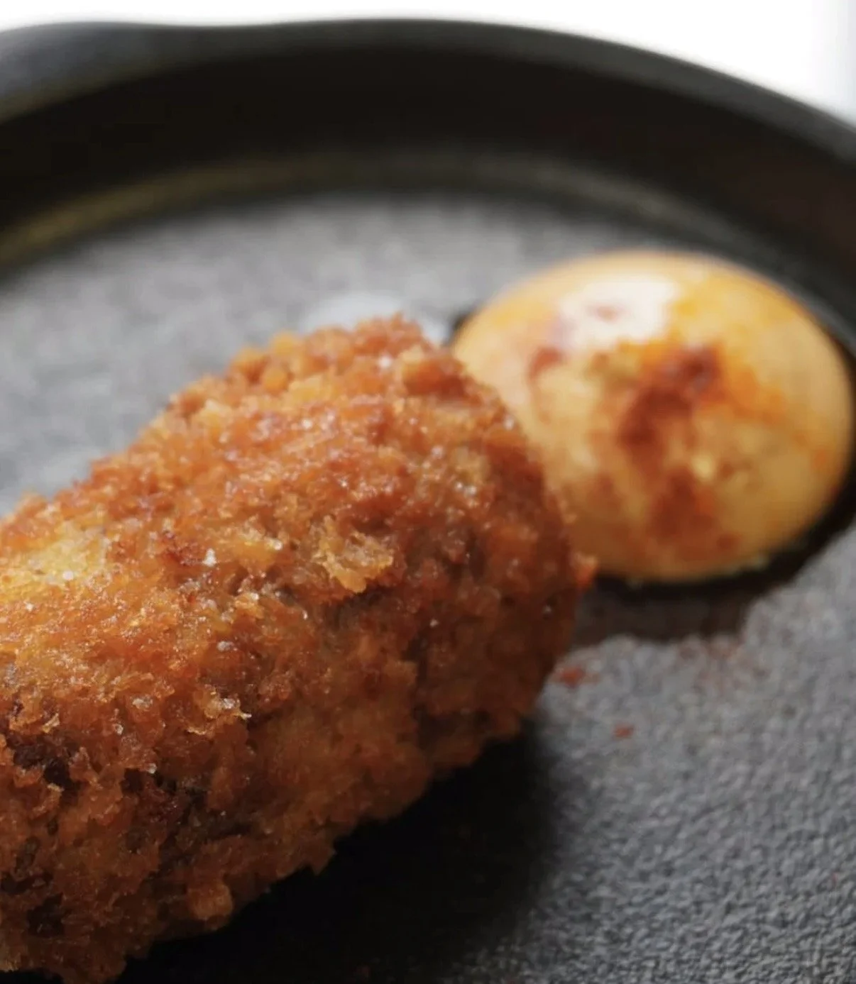 Close-up of a fried chicken drumstick with crispy breading and a boiled egg with paprika, on a black plate.