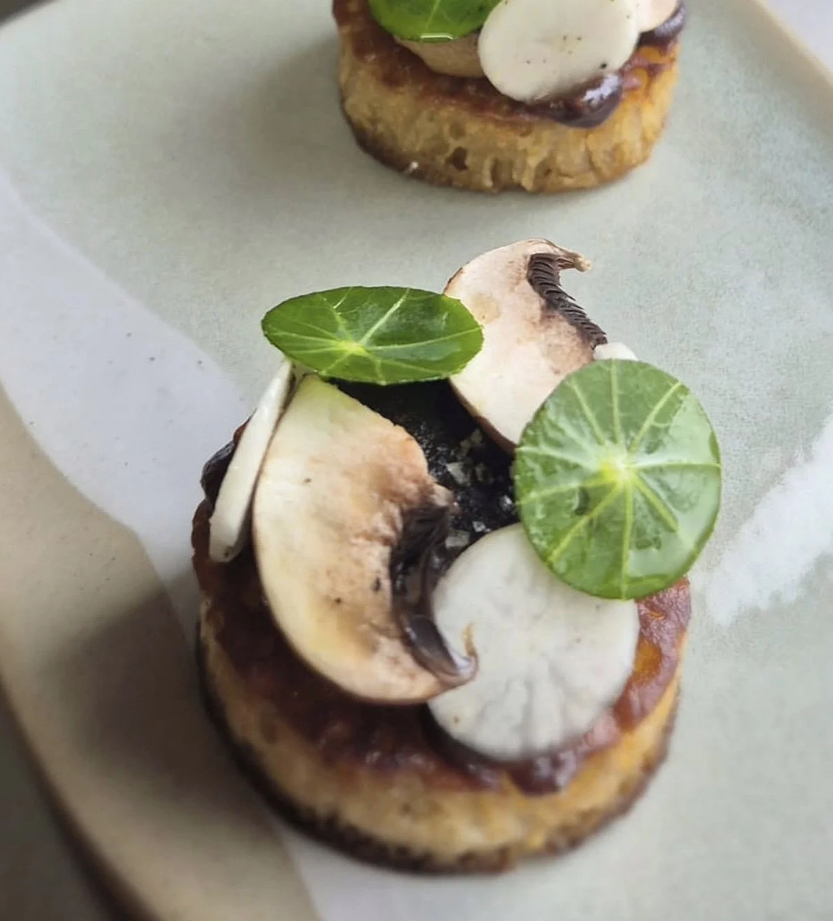 Close-up of a gourmet appetizer featuring toasted brown butter crumpet with slices of mushroom, dollops of white and dark sauces, and garnished with small green leaves.