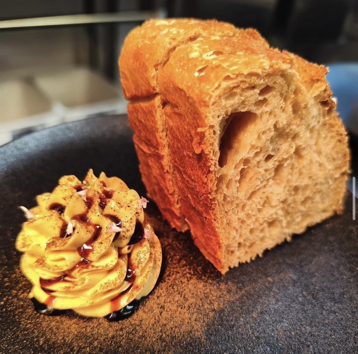 Close-up of a two thick slices of house-made sourdough on a dark plate, with a dollop of whipped butter topped with treacle and burnt leek powder.