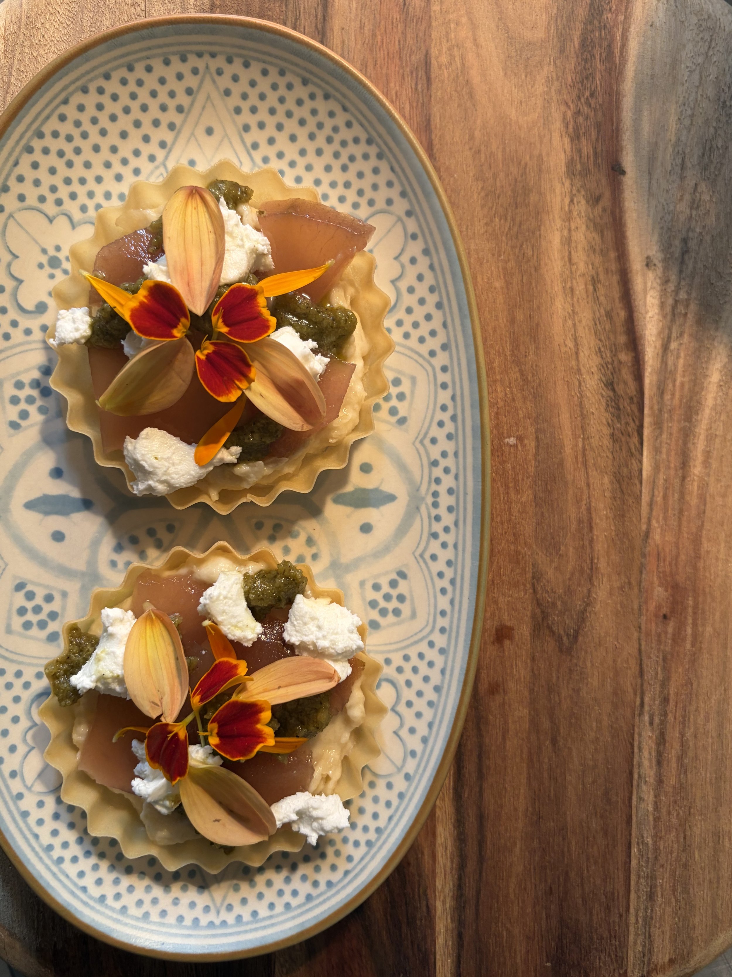 A decorative plate with small tart shells filled with fruit, topped with whipped cream and edible flower petals, on a wooden surface.