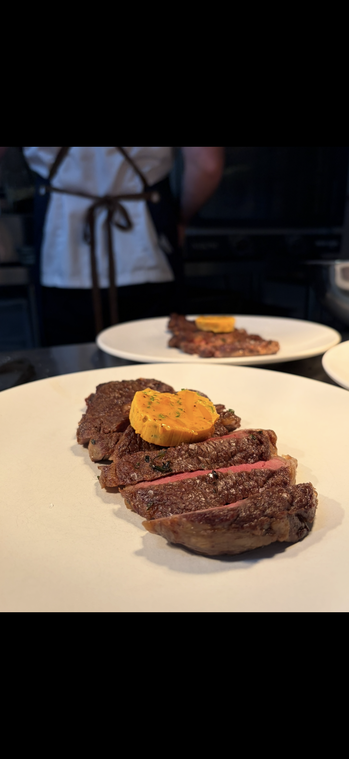 Close-up of sliced cooked steak topped with a yellow sauce on a white plate, with another plate of steak in the background