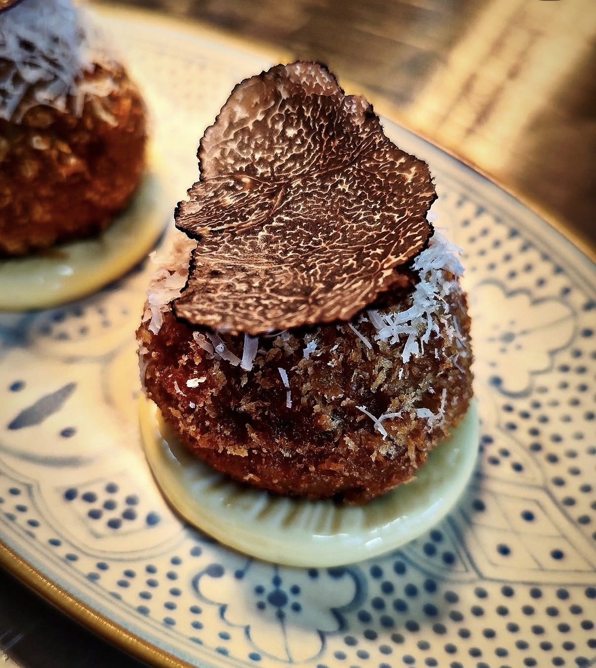 Close-up of a gourmet appetizer with a creamy yellow base, topped with a crispy, breaded meatball, shredded cheese, and a thin slice of black truffle on top, served on a decorative plate.