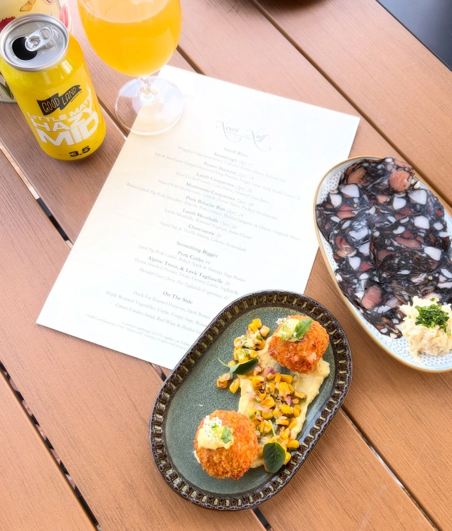 A table set with a menu, a glass of orange juice, a yellow soda can, and various dishes of food including a bowl of dessert with chocolate topping, a plate of mashed potatoes with a croquette and corn, and a plate with two croquettes and a small sala