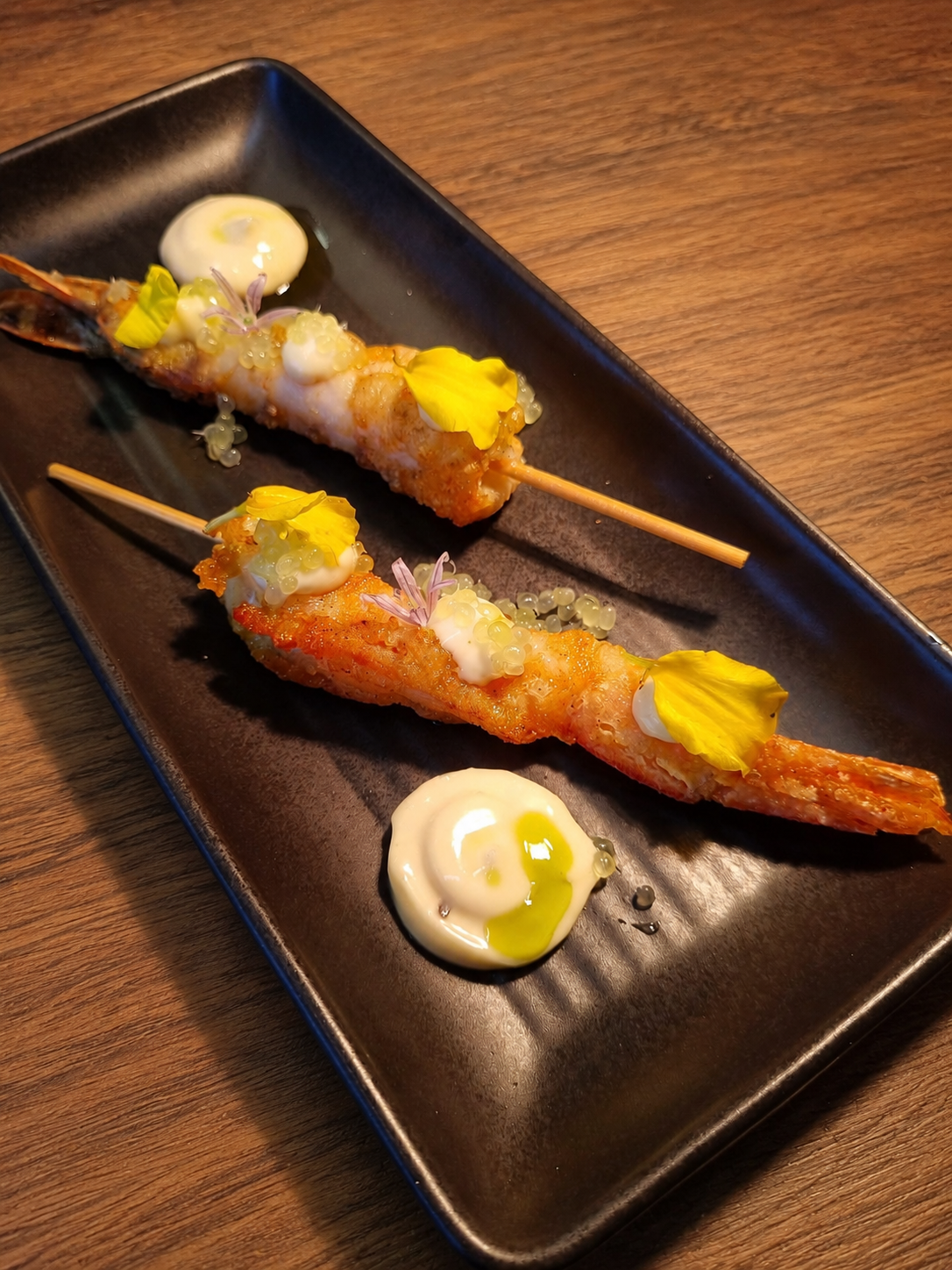 Two skewers of grilled King Prawns garnished with edible flowers and small dollops of finger lime aioli, served on a black rectangular plate with additional sauce dollops.