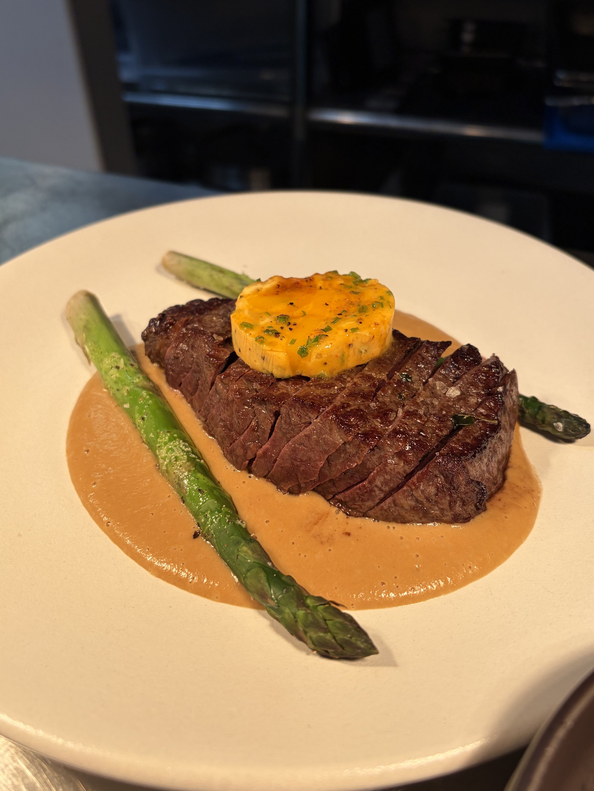 A plate with a pan seared scotch fillet with a round slice of gascony butter, accompanied by two stalks of grilled asparagus and a creamy pepper sauce underneath the steak.