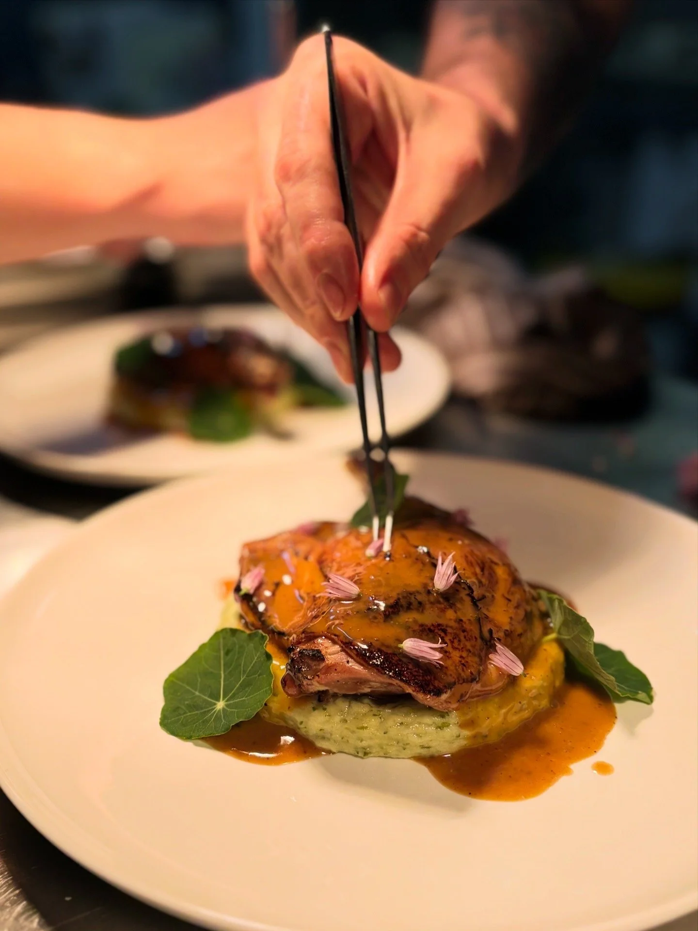 A plated gourmet dish featuring a piece of roasted meat topped with sauce, garnished with pink flower petals and green leaves, with additional plates of similar dishes in the background.