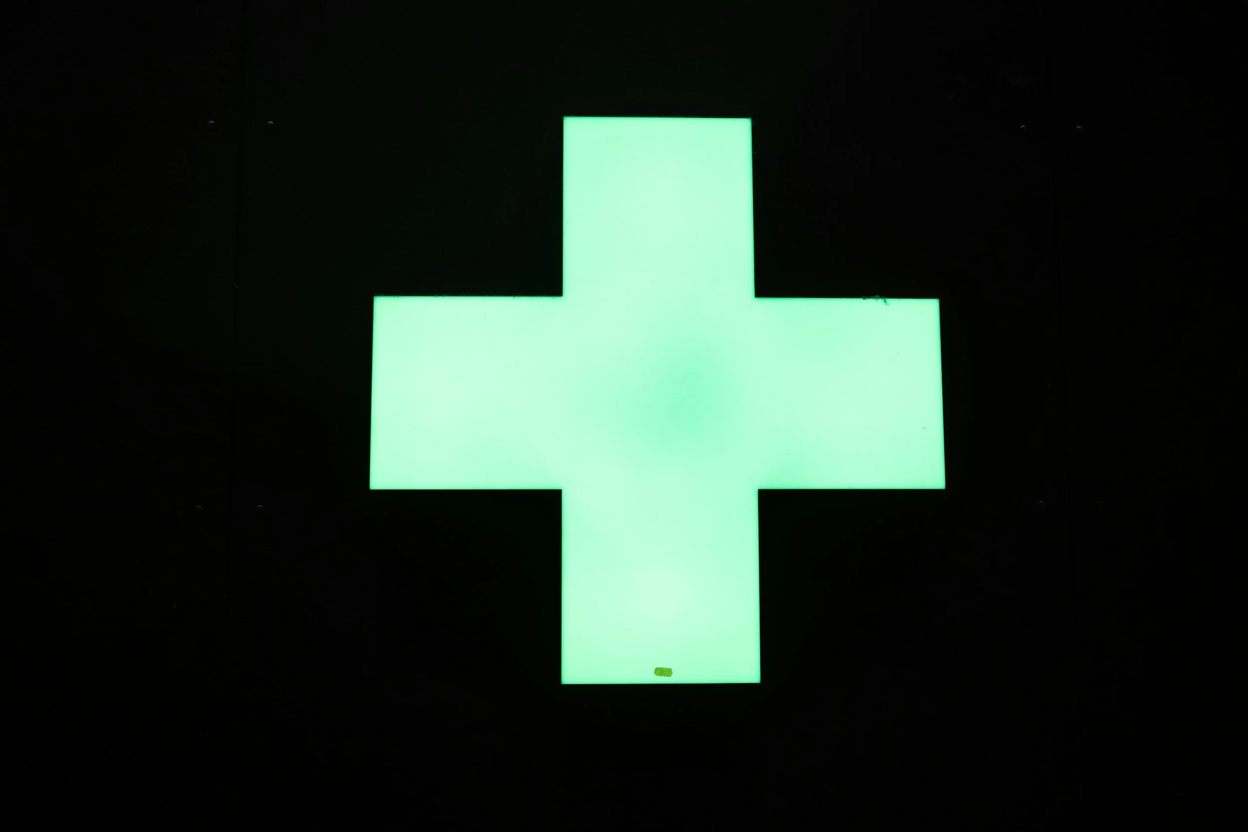 Green illuminated cross symbol on a dark background, representing a pharmacy or medical facility.