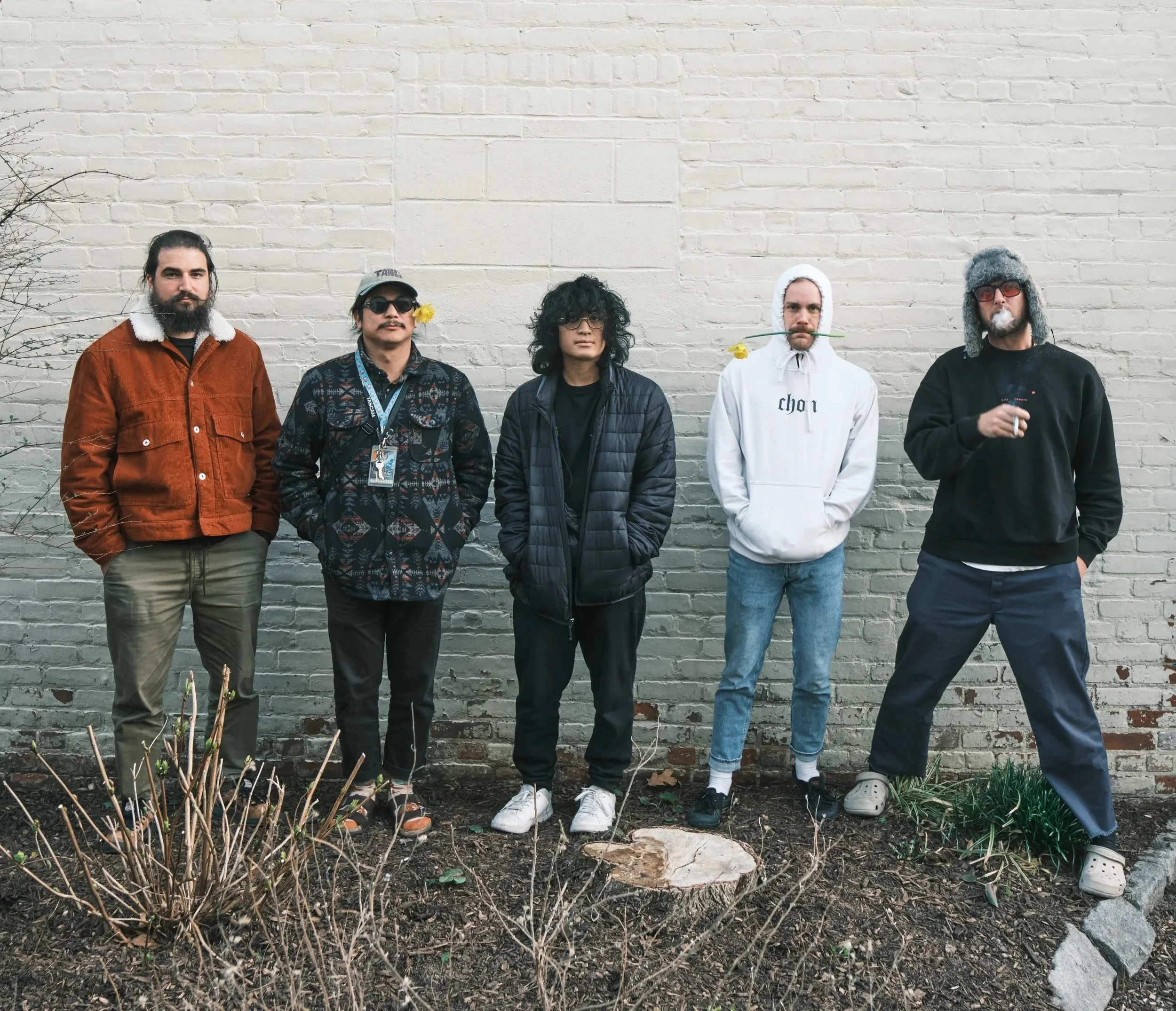 Five men standing in front of a beige brick wall, dressed in casual winter clothing, with some wearing hats or glasses, and one holding a cigarette.
