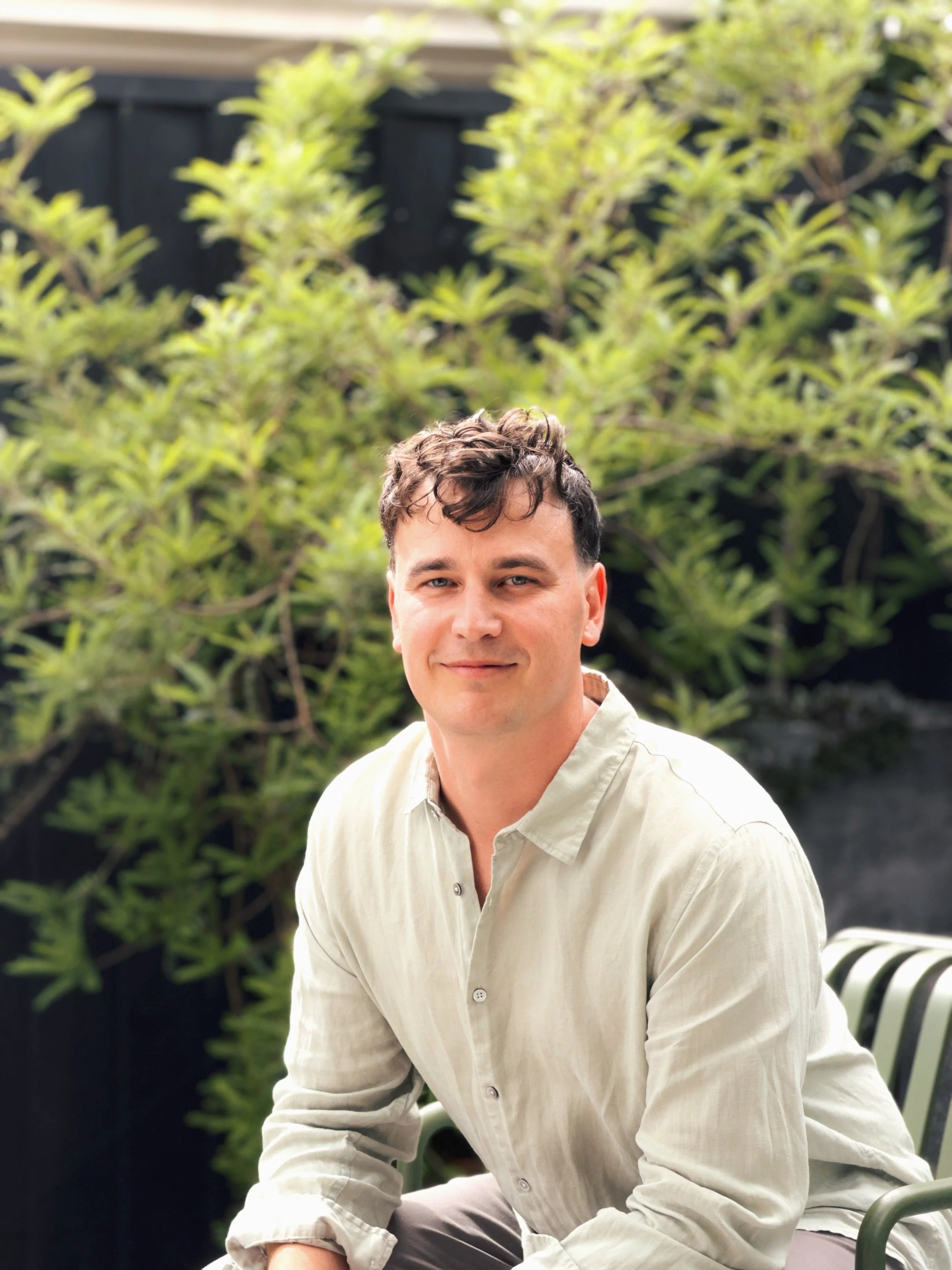 Nathan smiling warmly at the camera, seated outdoors in a garden setting, wearing a light linen button-up shirt with lush green foliage in the background.