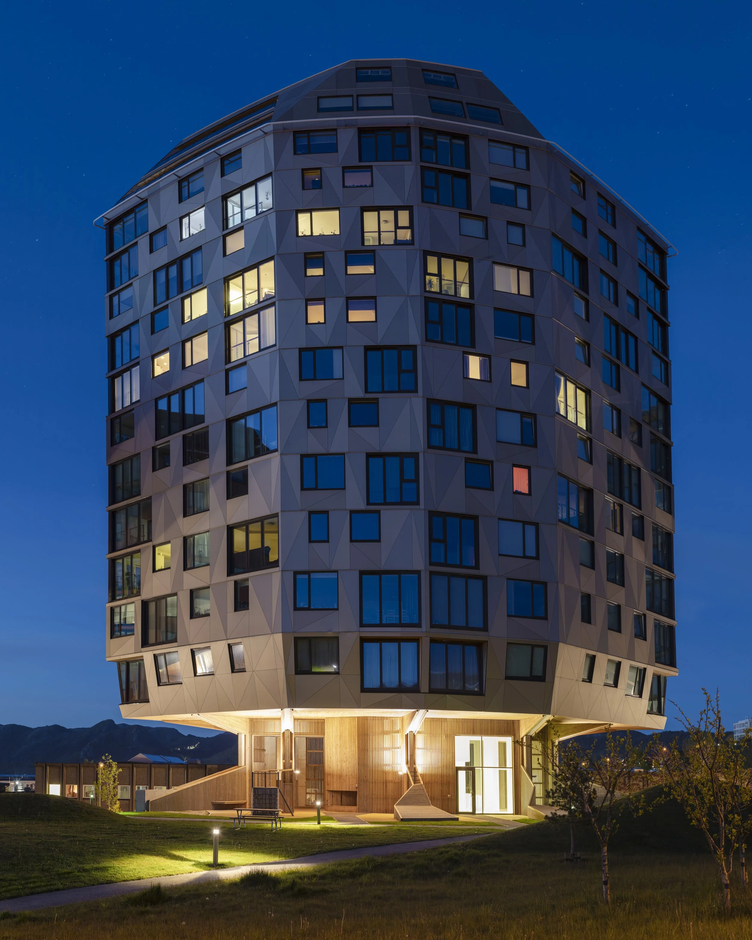Modern multi-story building with a unique geometric facade, illuminated windows, and a well-lit ground level, set against a twilight sky.