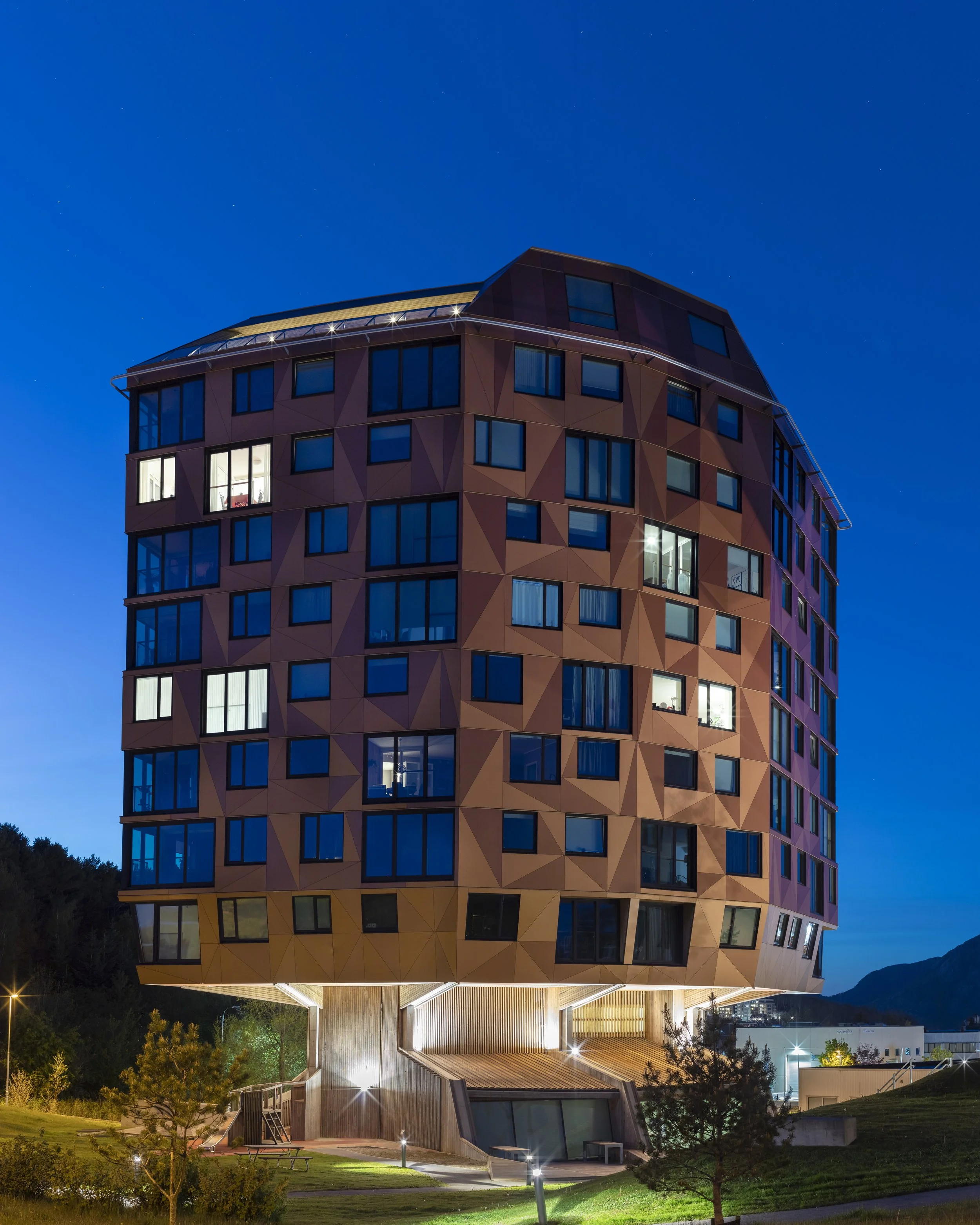 Unique multi-story building with an asymmetrical, geometric copper-colored facade and many windows, illuminated at night.