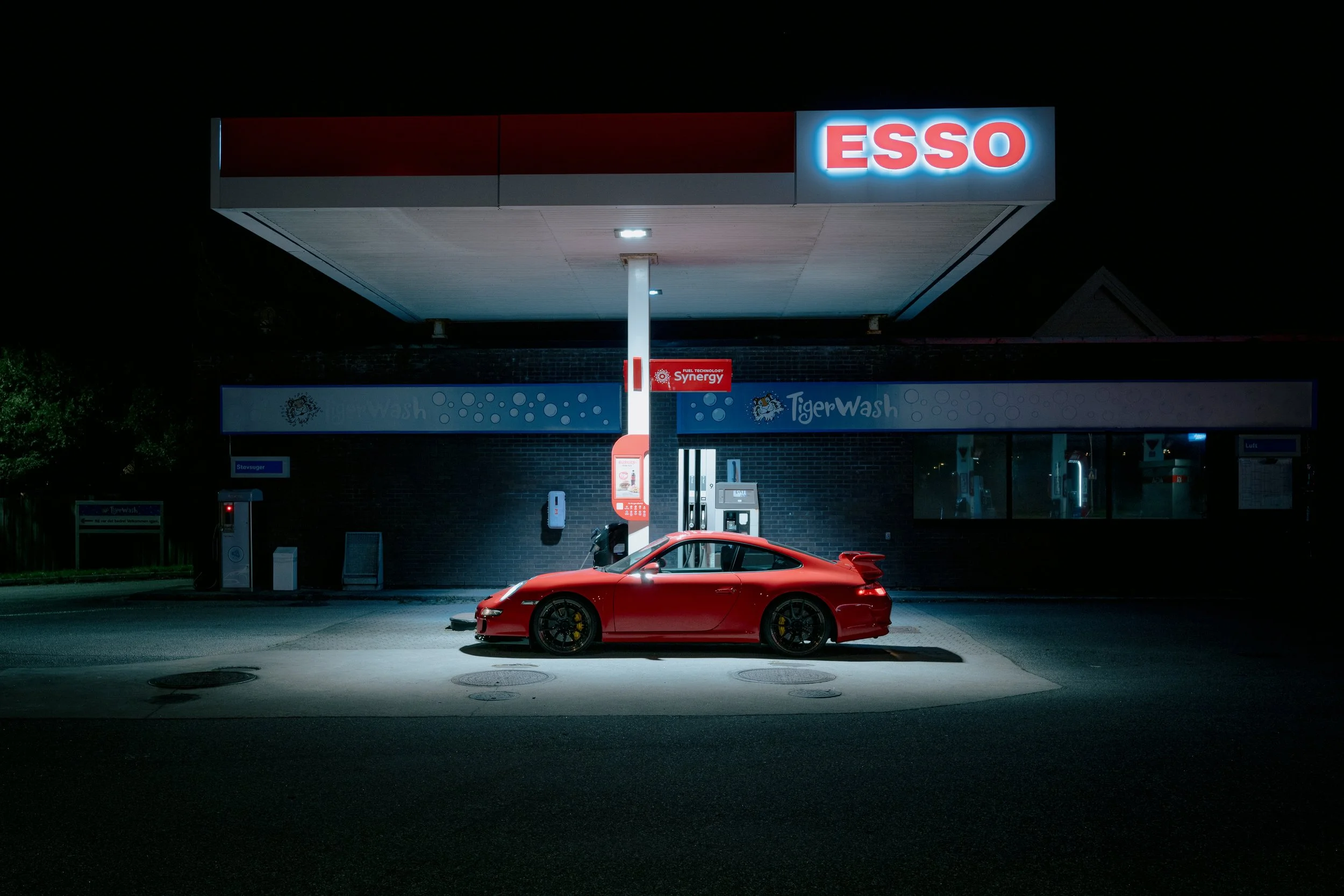 Red sports car parked at Esso gas station at night with illuminated signage and pump area.