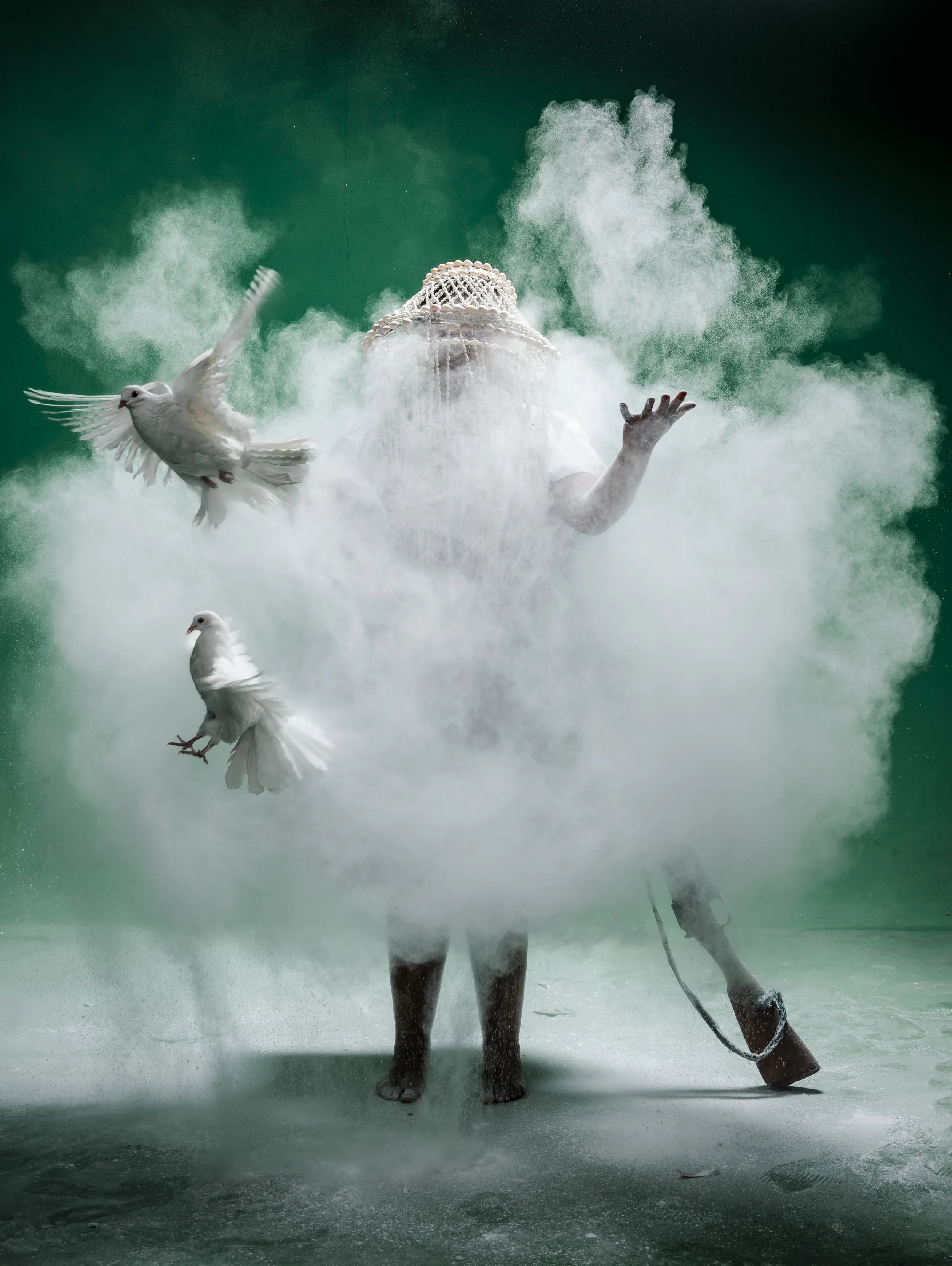 A person with a woven hat obscuring their face surrounded by white pigeons and a green smoky atmosphere, standing with one arm raised.