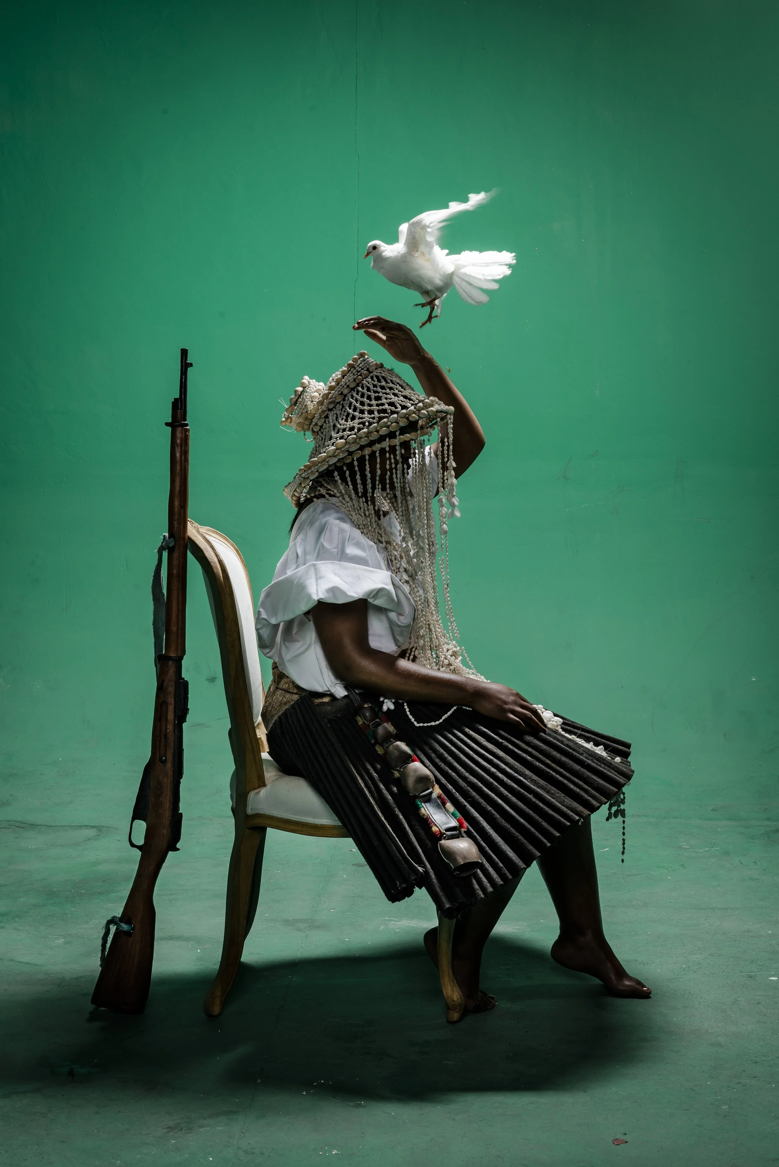 A person wearing traditional clothing and a beaded headpiece sitting on a chair with a rifle leaning against it, a white dove is flying above their hand, set against a plain green background.