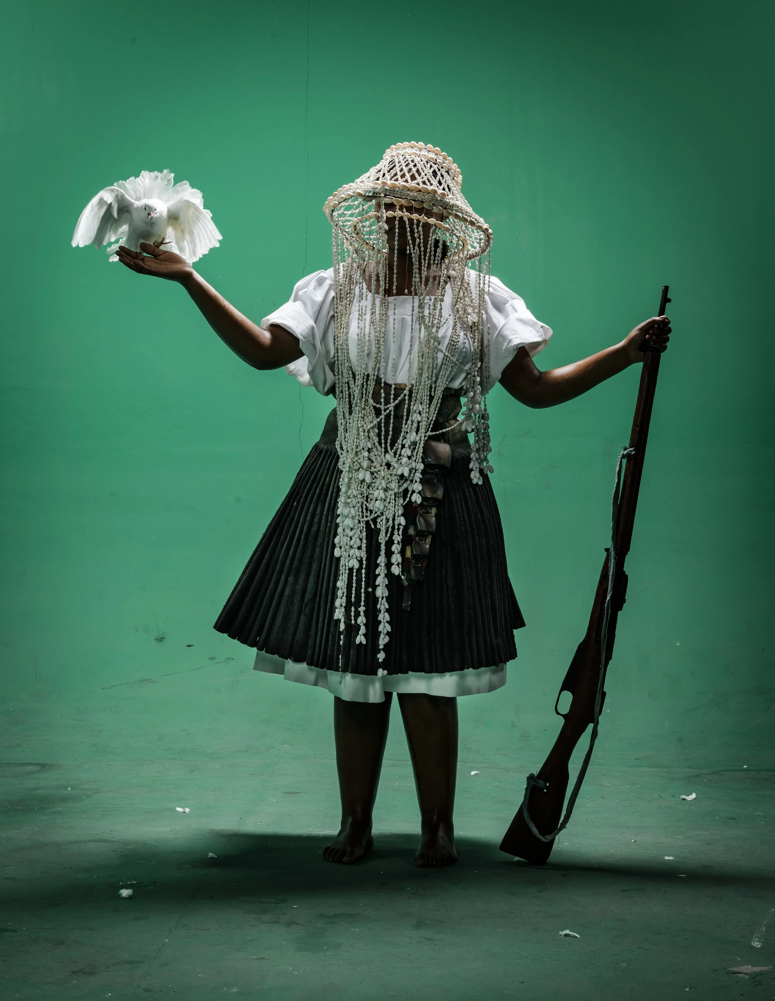 Person in traditional attire standing barefoot against a green background, holding a white dove in one hand and a rifle in the other, wearing a hat decorated with shells or beads, and adorned with strings of beads and shells.