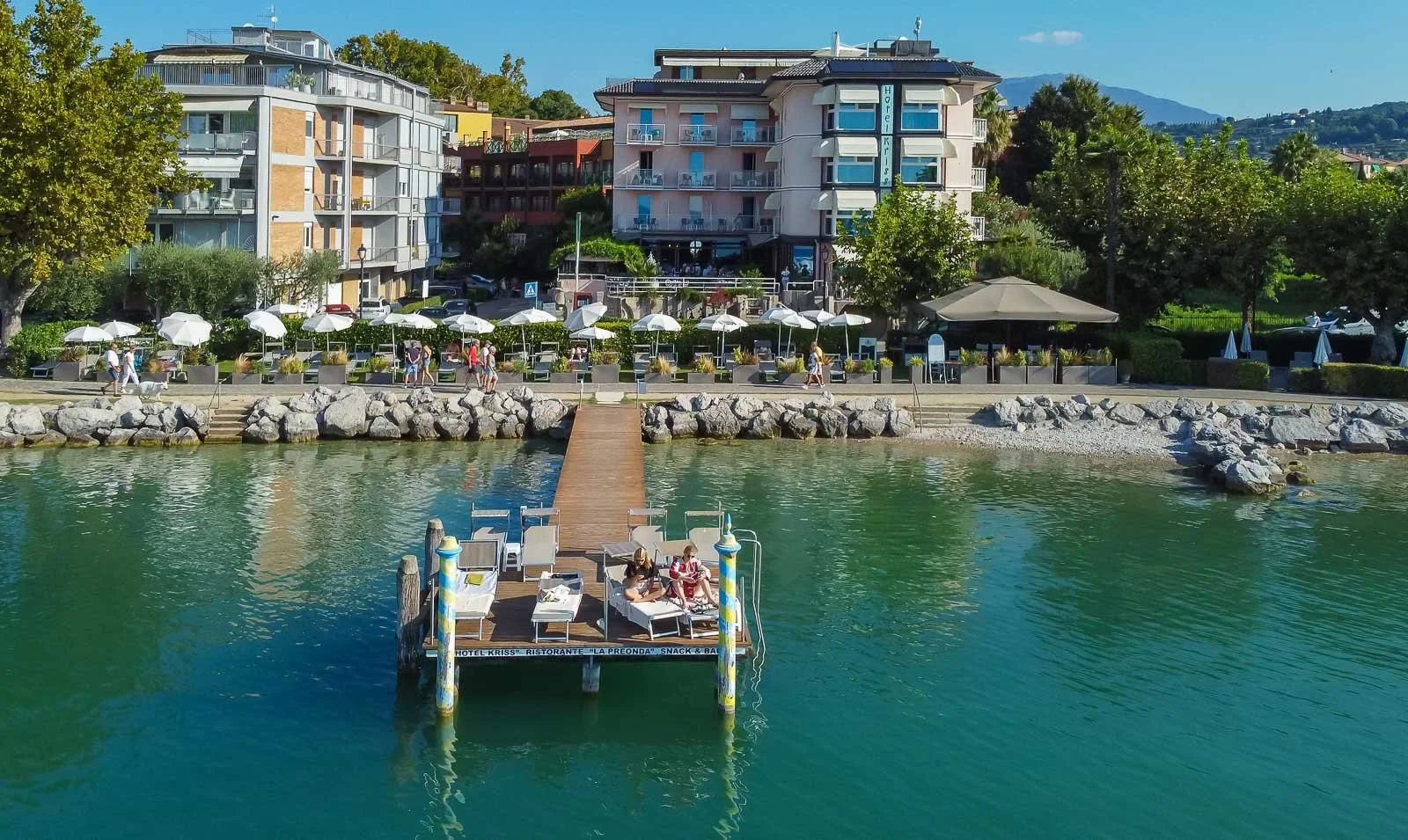 Vista di un lungolago con un pontile in legno e sedie a sdraio, sullo sfondo edifici residenziali e alberi, con alcune persone che passeggiano e si rilassano.