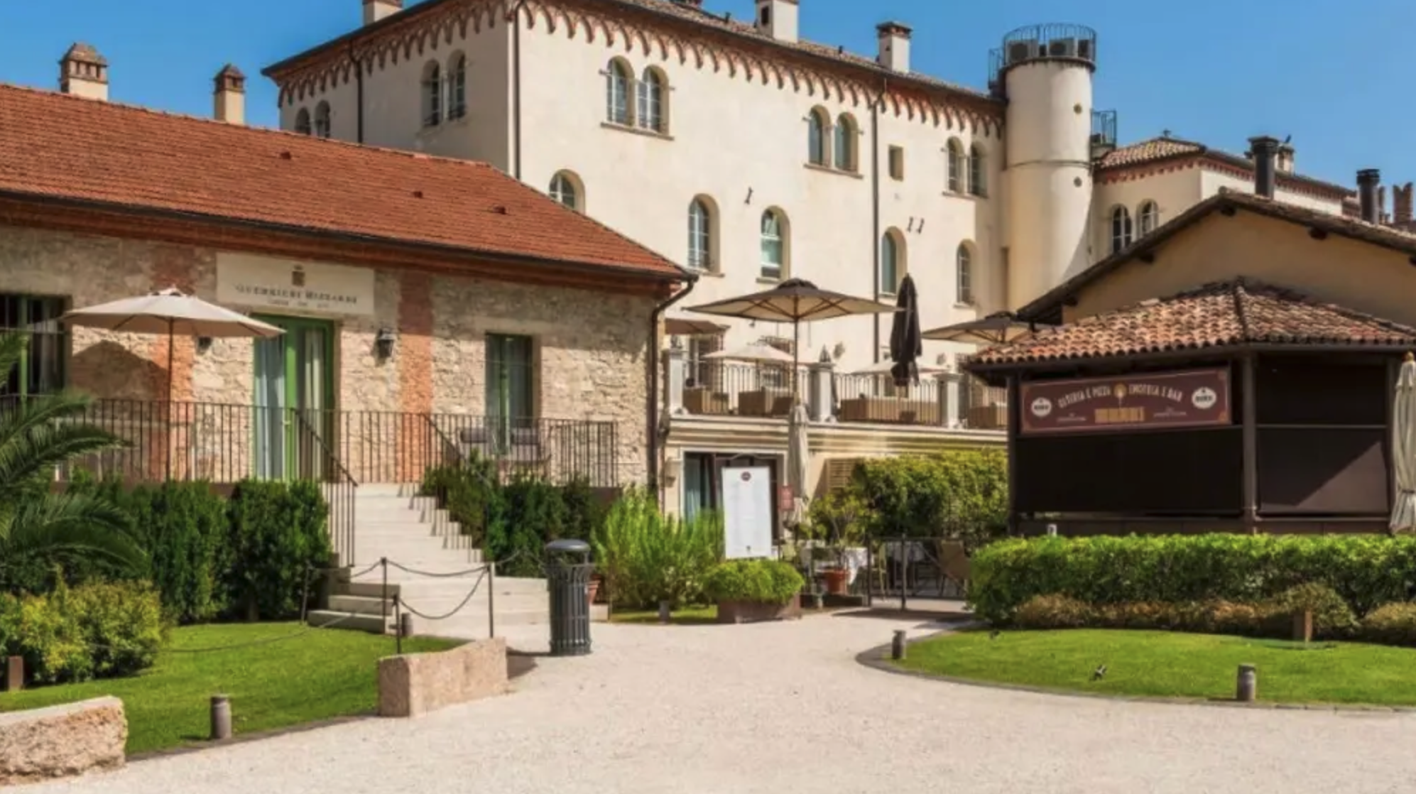 Uncastello medievale con muri bianchi e torri, circondato da un giardino ben curato con siepi e piante, con un'area esterna con ombrelloni e sedie.