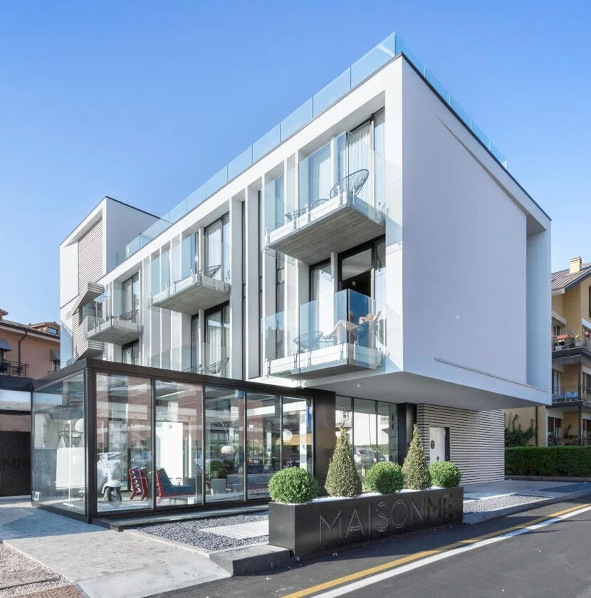 Edificio moderno con balconi in vetro e un'area lounge esterna in vetro, alberi decorativi e il nome 'MAISONNE' nella posizione frontale.