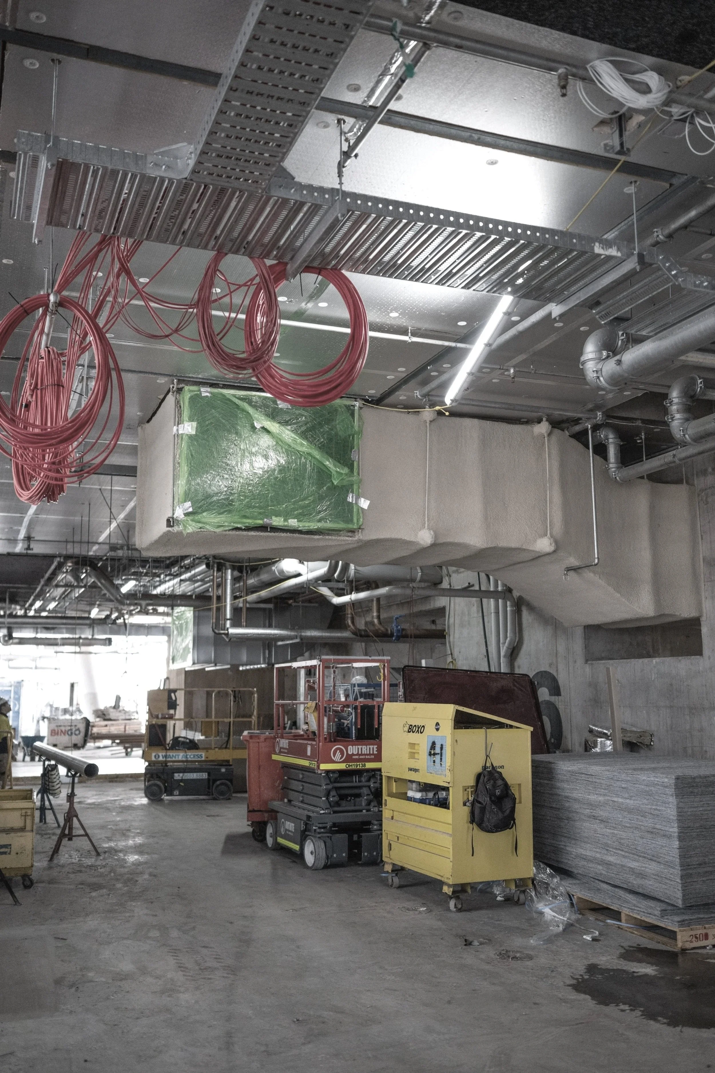 Construction site with ductwork, electrical wiring, and construction equipment including scissor lifts and material stacks.