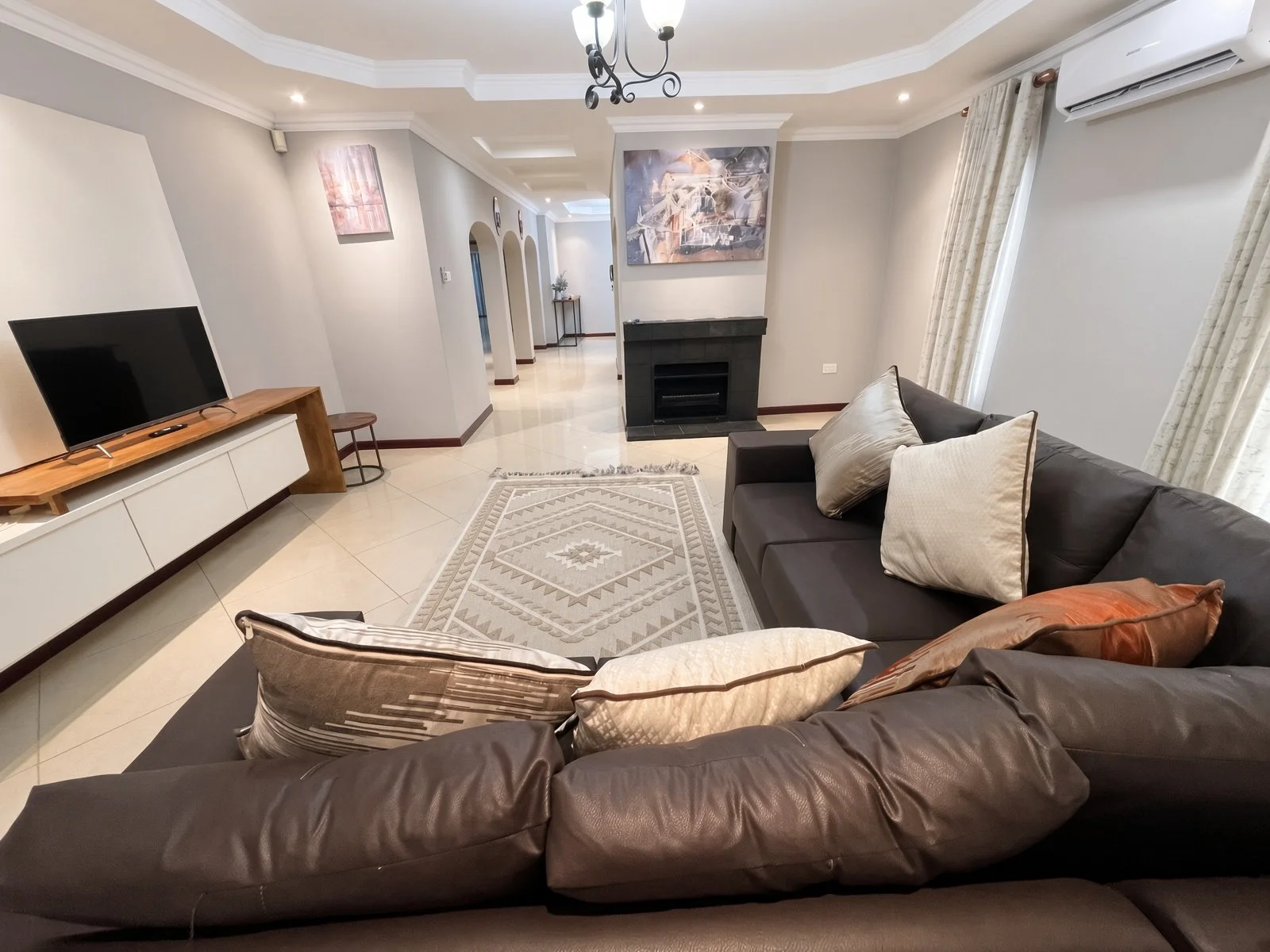 Open-plan lounge with fireplace and view through to the hallway in this fully furnished 5-bedroom house for rent in Phakalane. Nestgeeks.