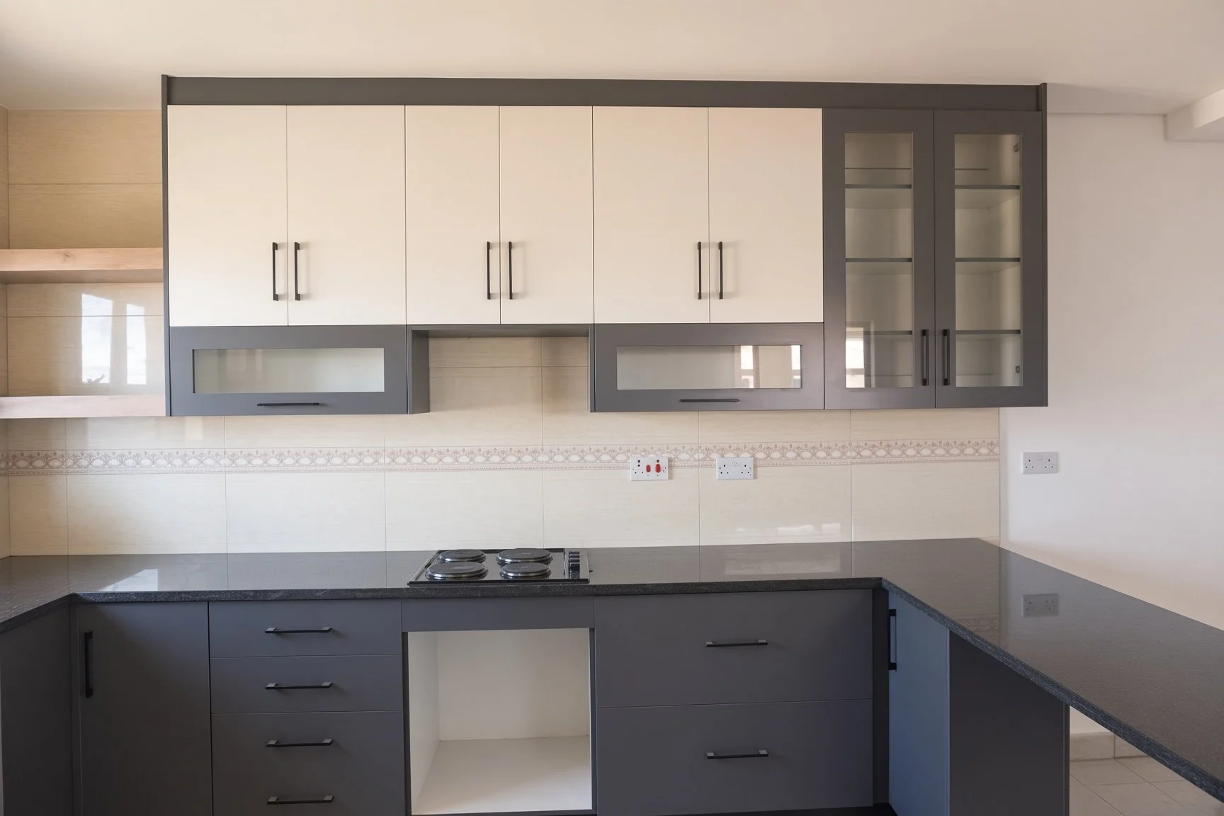 Fitted open-plan kitchen with cream and charcoal cabinets, glass-front display units, black granite worktops and two-plate hob in the 180 sqm 3-bedroom unit for rent, gated estate Gaborone North. Nestgeeks.