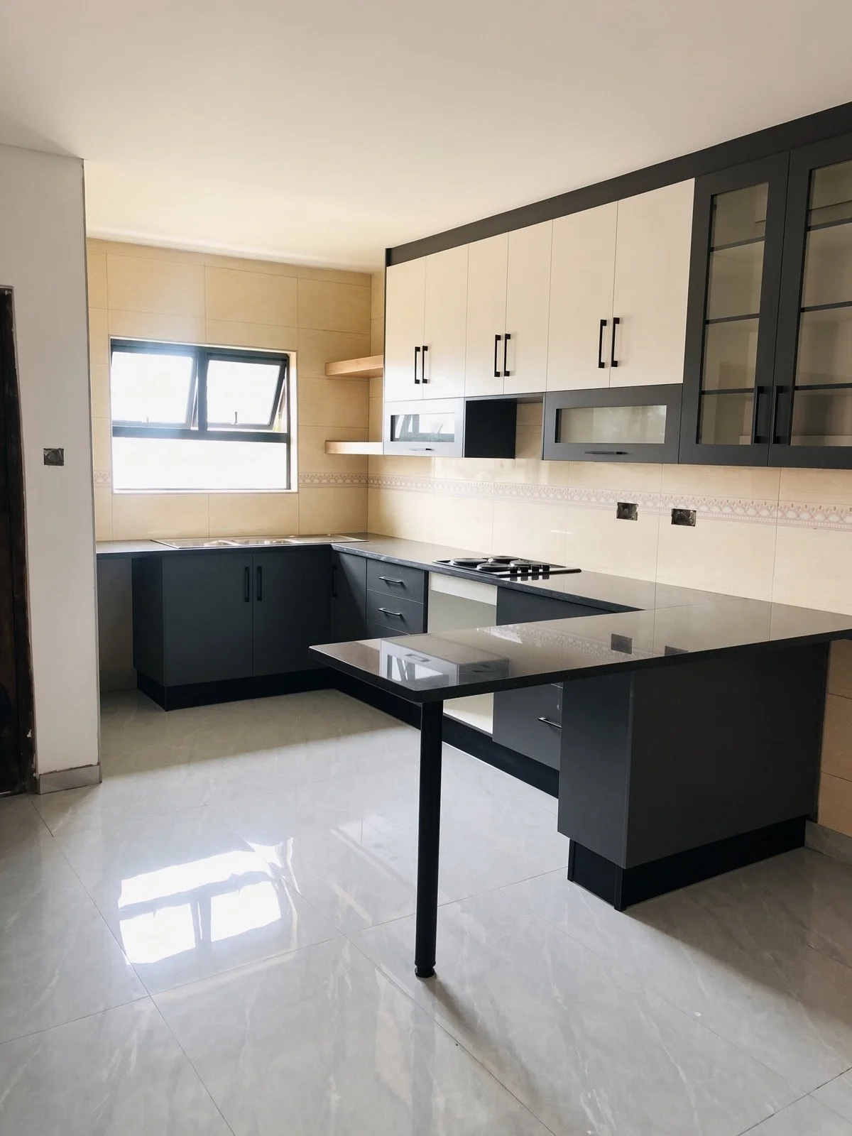 Open-plan kitchen with L-shaped granite counters, breakfast bar, fitted grey and cream units and window in the modern 3-bedroom rental unit, gated estate Gaborone North. Listed by Nestgeeks.