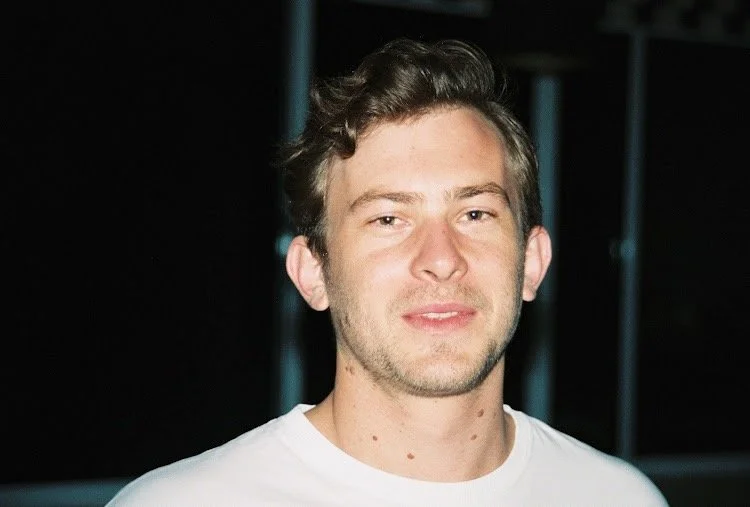 A young man with light skin, short brown hair, and blue eyes, standing outdoors at night, wearing a white T-shirt.