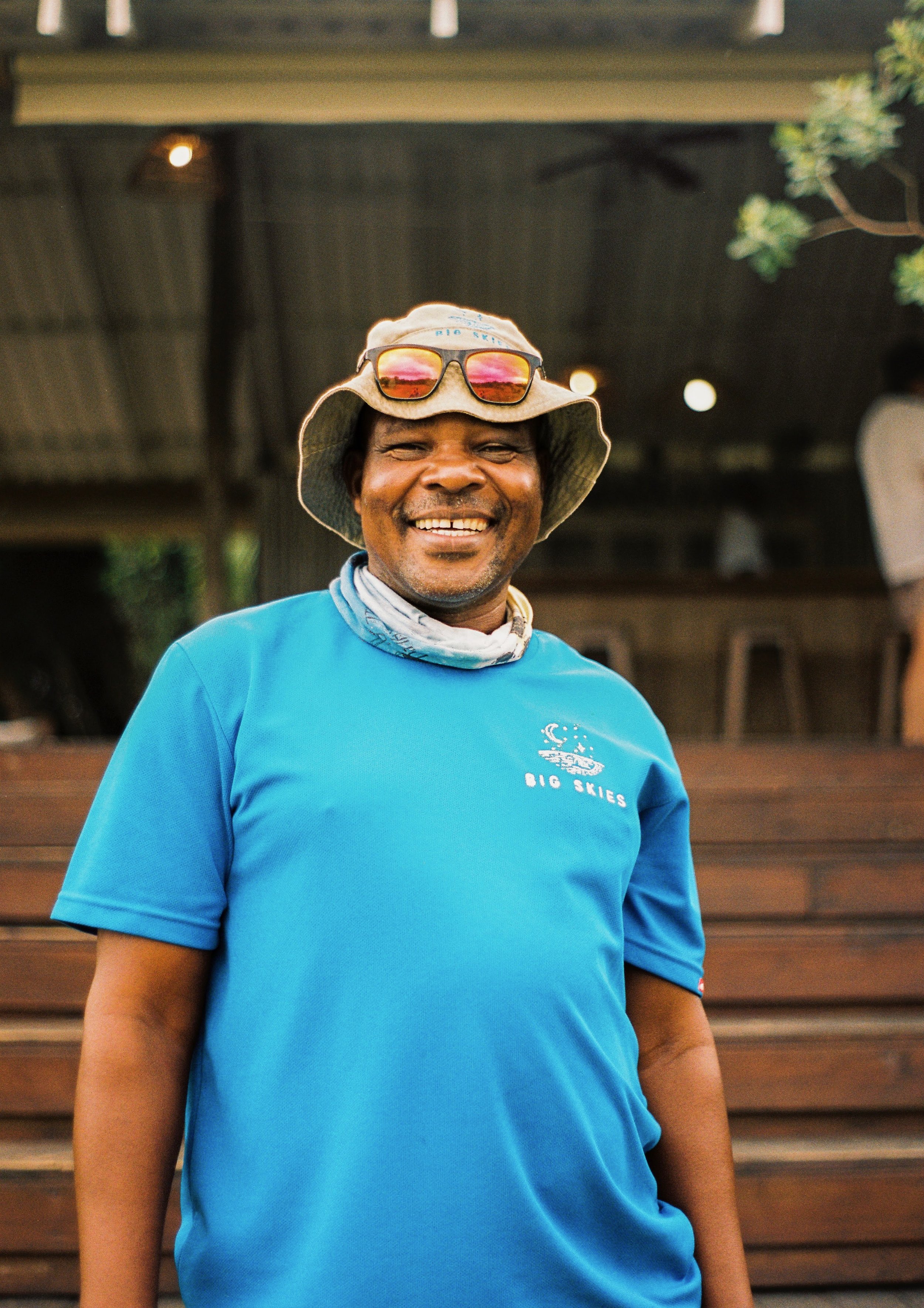 A man smiling while wearing a blue Big Skies t-shirt, a wide-brimmed hat with sunglasses on top, and a scarf around his neck, standing outdoors with a wooden background.